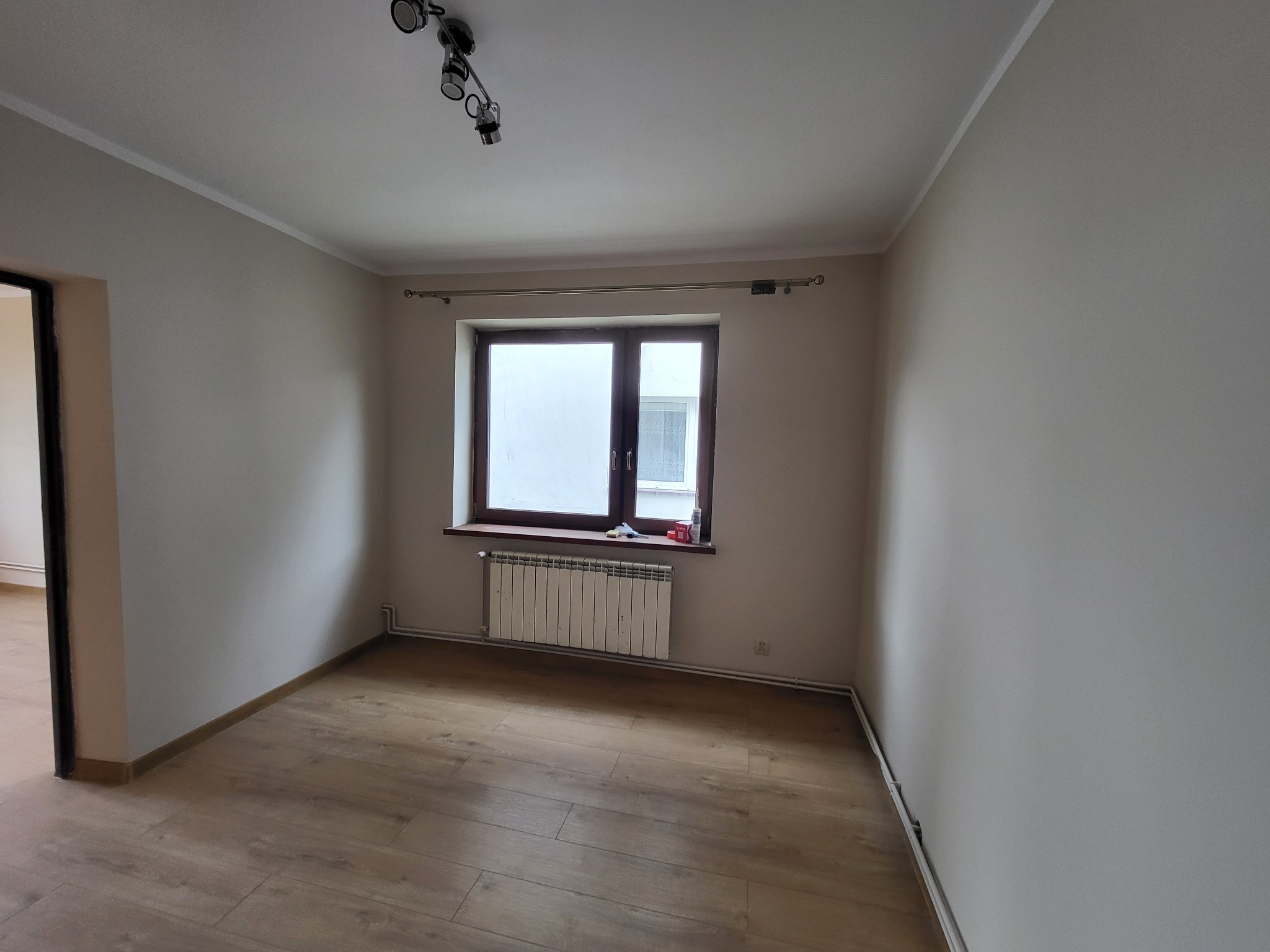 Pusty pokój po remoncie z drewnopodobną podłogą, świeżo pomalowanymi ścianami w odcieniach beżu, grzejnikiem pod oknem i widoczną listwą przypodłogową.