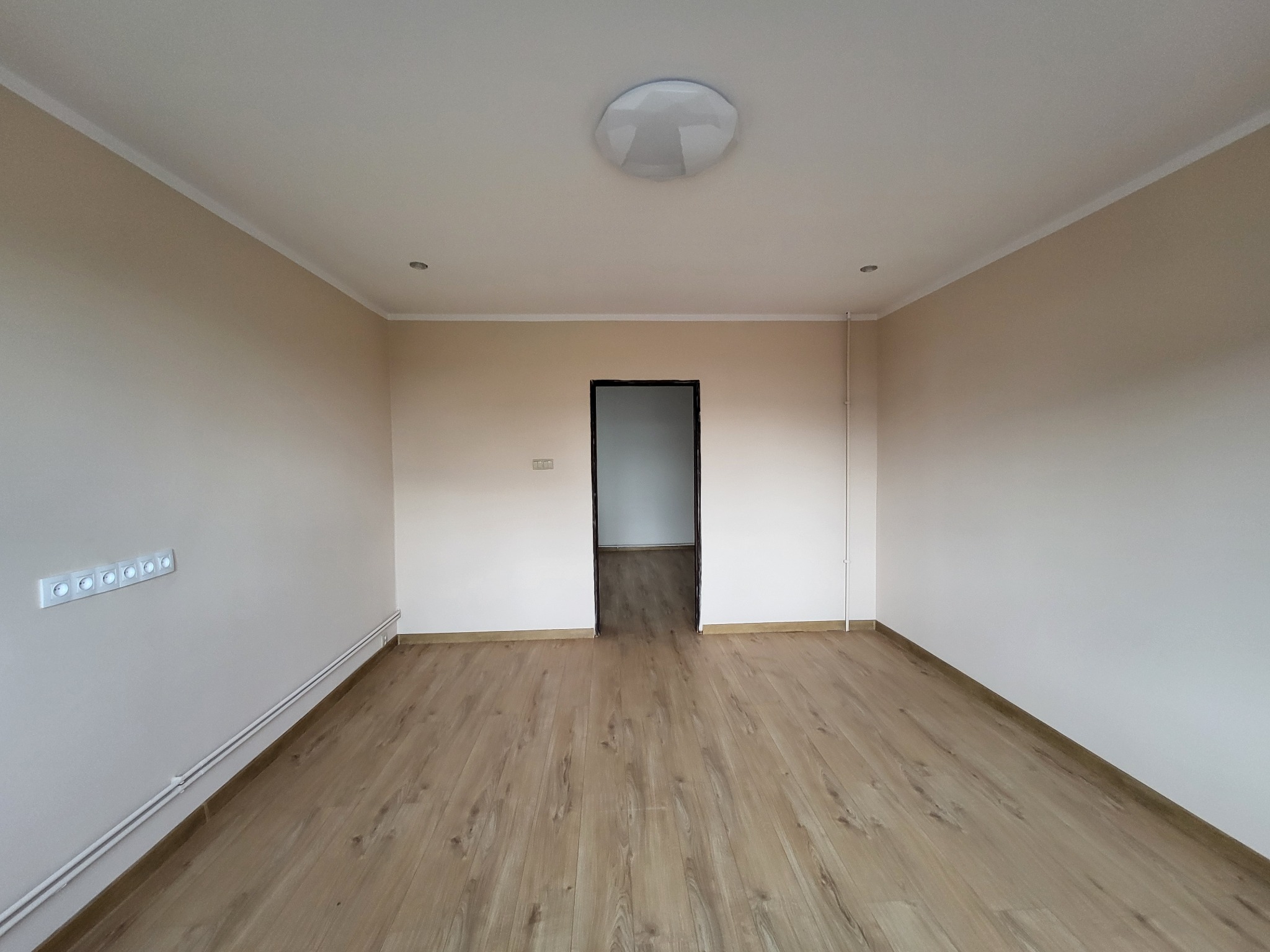 Pomalowane na beżowo puste pomieszczenie z ułożonymi panelami podłogowymi, widoczne gniazdka elektryczne i lampa sufitowa.