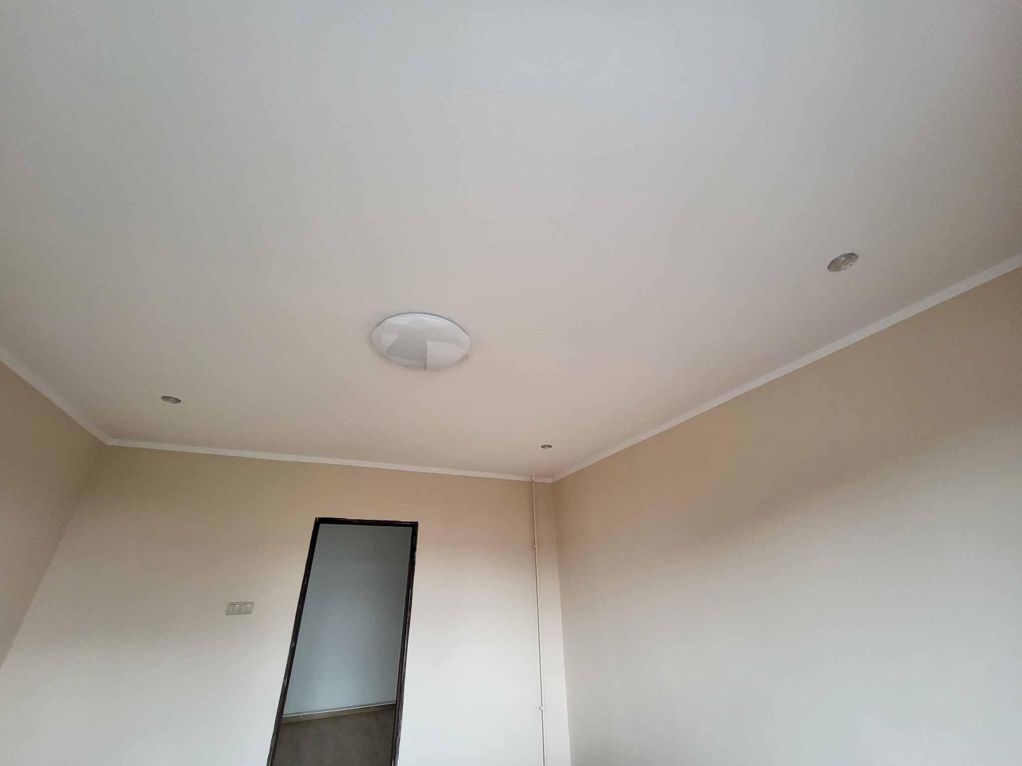 Pomalowane na beżowo ściany i biały sufit z zamontowanym okrągłym plafonem i dwoma małymi punktowymi lampkami halogenowymi, widoczne listwy przypodłogowe i ościeżnica drzwi.