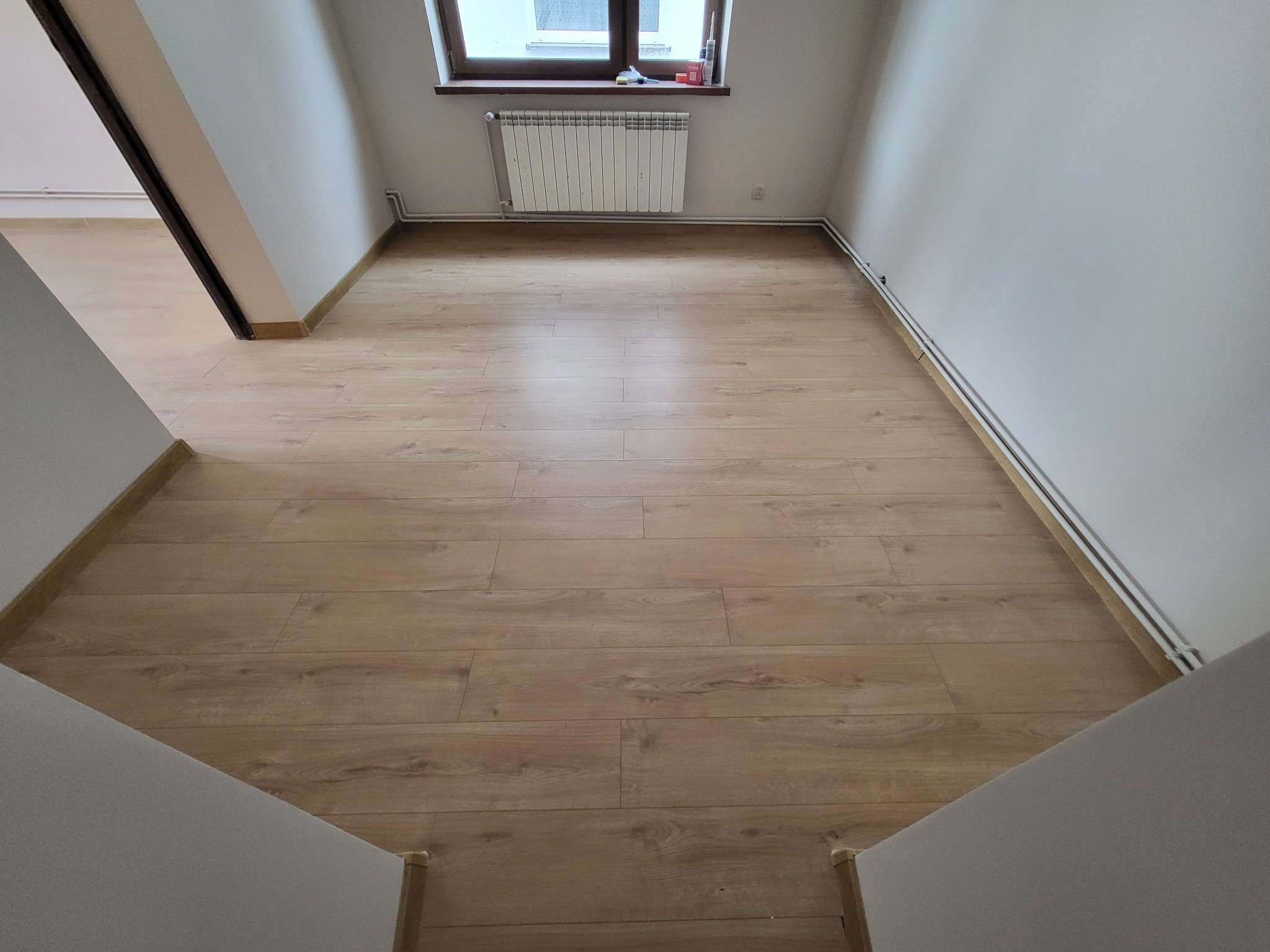Jasne panele podłogowe w nowym pokoju na poddaszu, widoczny grzejnik pod oknem i białe ściany.