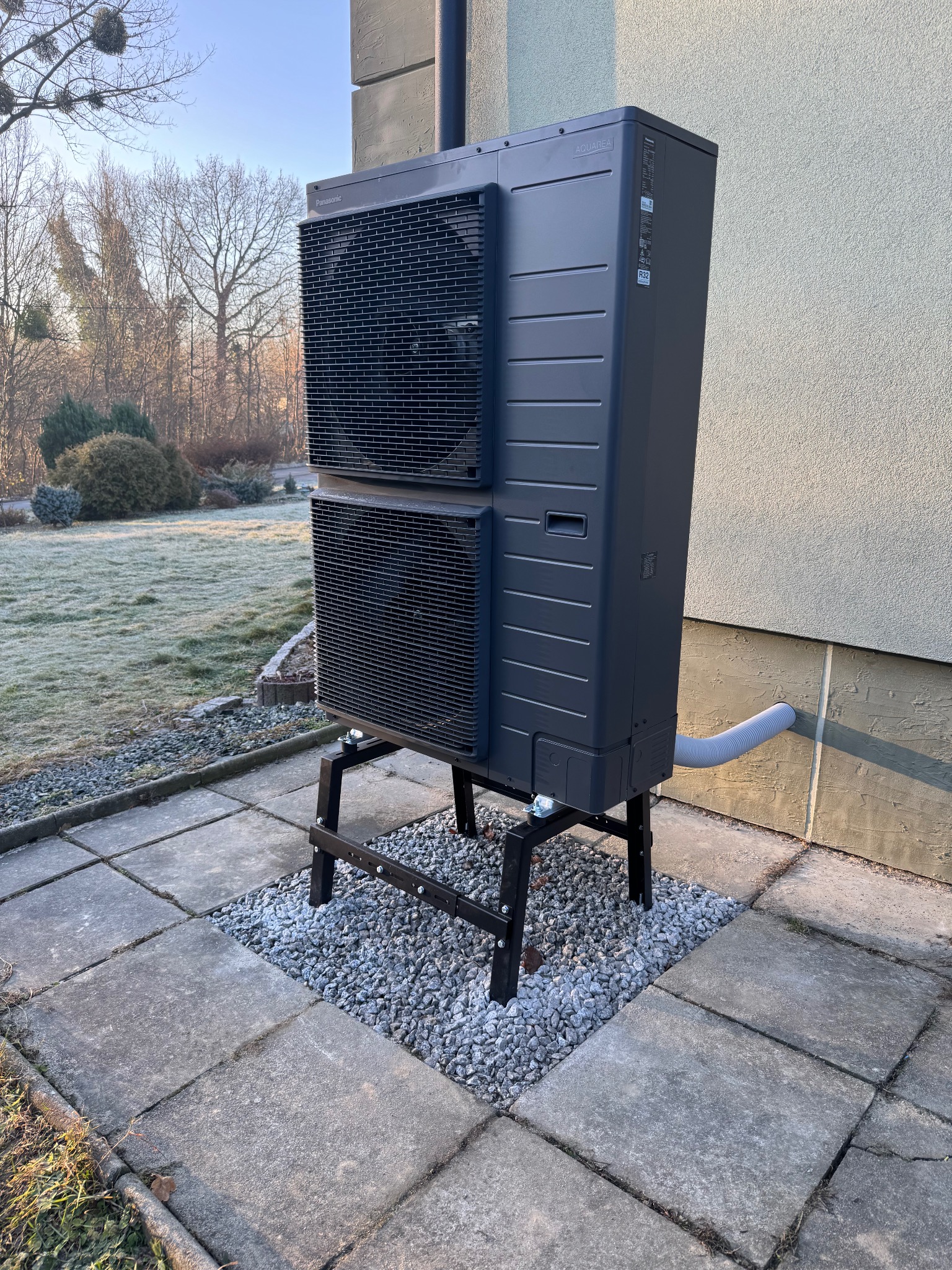 Zewnętrzna jednostka pompy ciepła Panasonic Aquarea zamontowana na czarnej, metalowej podstawie na tle ogrodu z oszronioną trawą.