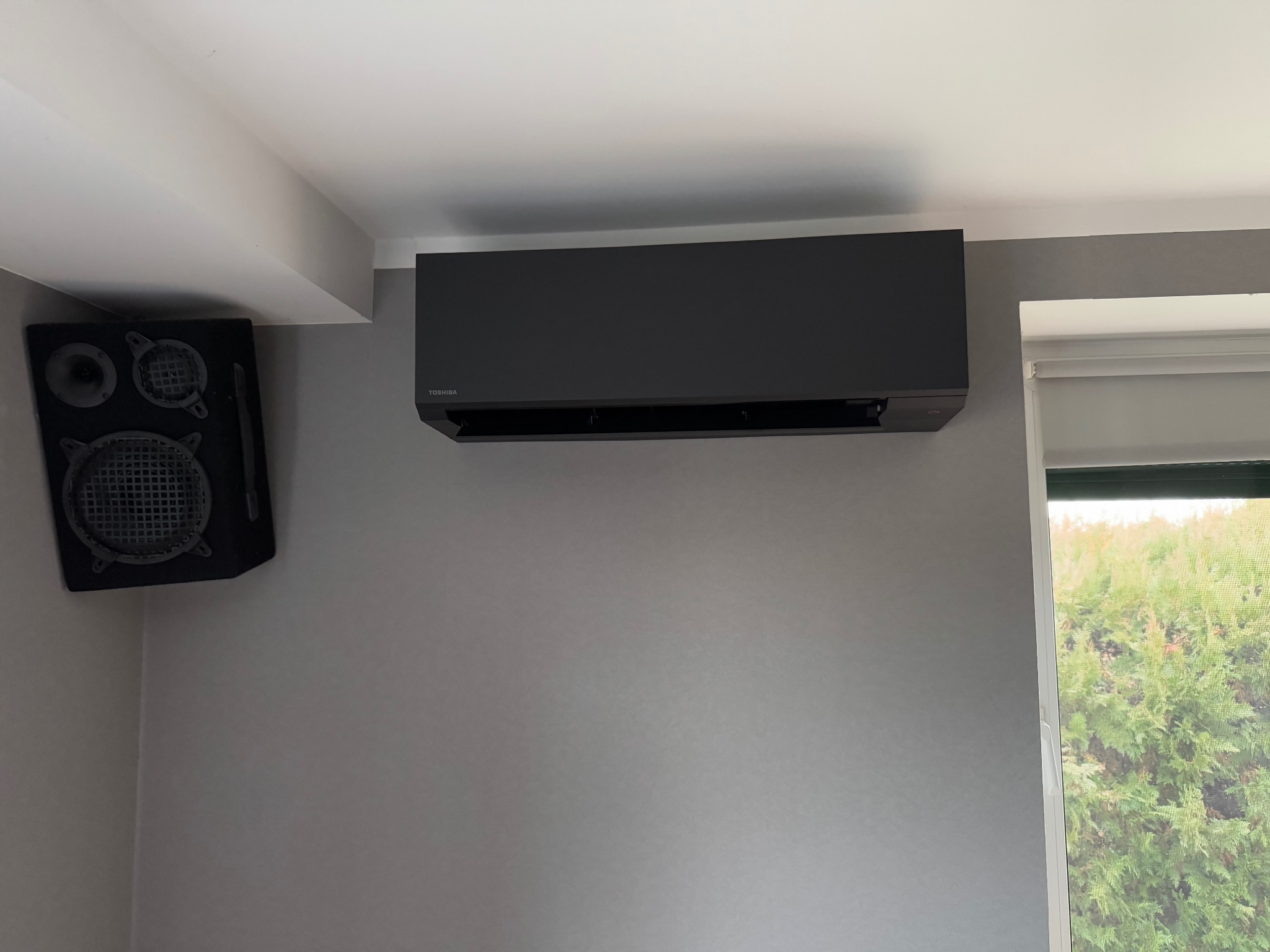 Czarna klimatyzacja Toshiba zamontowana na szarej ścianie w pokoju, obok głośnik i okno z widokiem na zieleń.