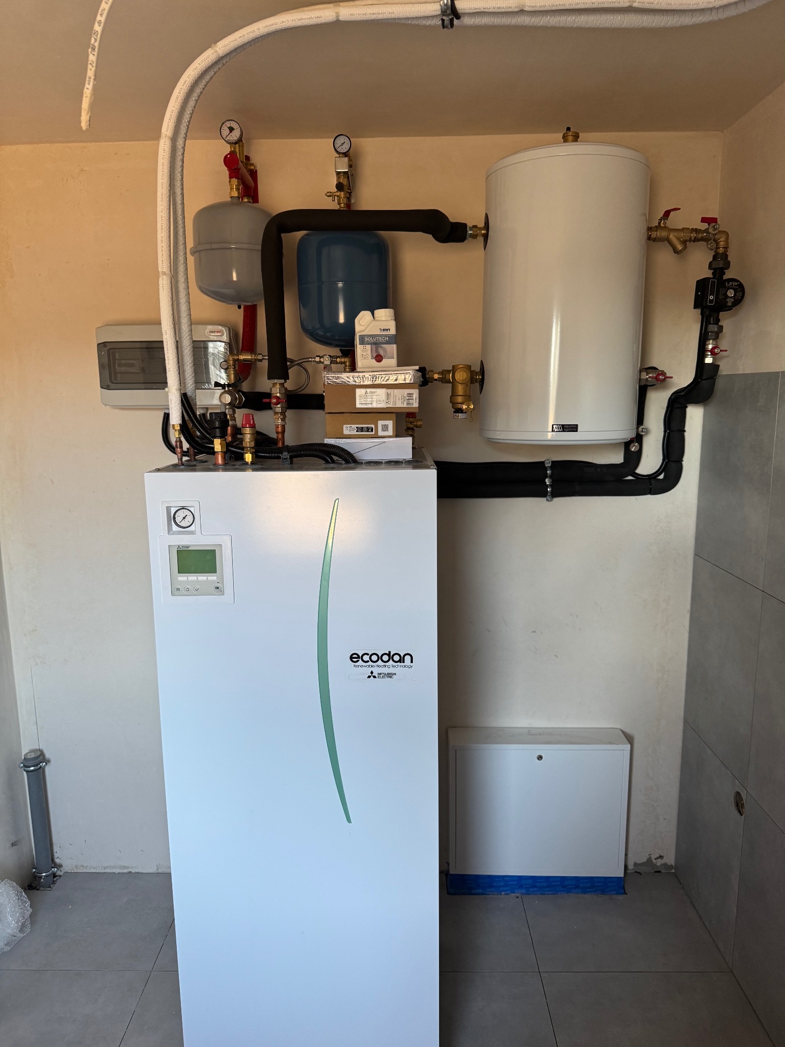 Biała pompa ciepła Mitsubishi Ecodan z podłączonymi rurami, zbiornikiem wyrównawczym i zasobnikiem CWU w kotłowni z szarymi płytkami