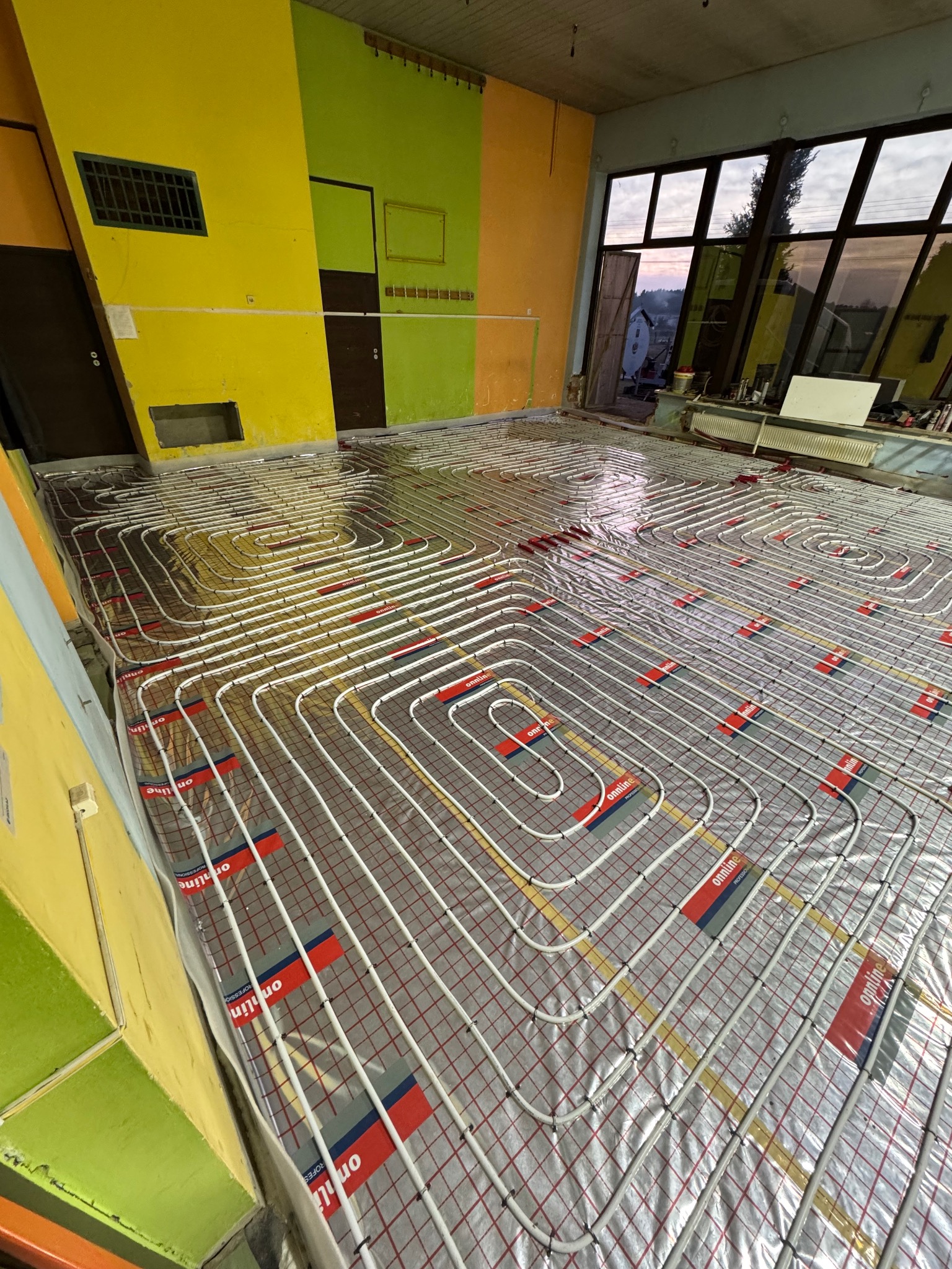 Rozłożone rury ogrzewania podłogowego na folii izolacyjnej z czerwono-szarymi oznaczeniami 'onntline PROFESSIONAL', widok na wielobarwne ściany i okno z widokiem na zachodzące słońce.