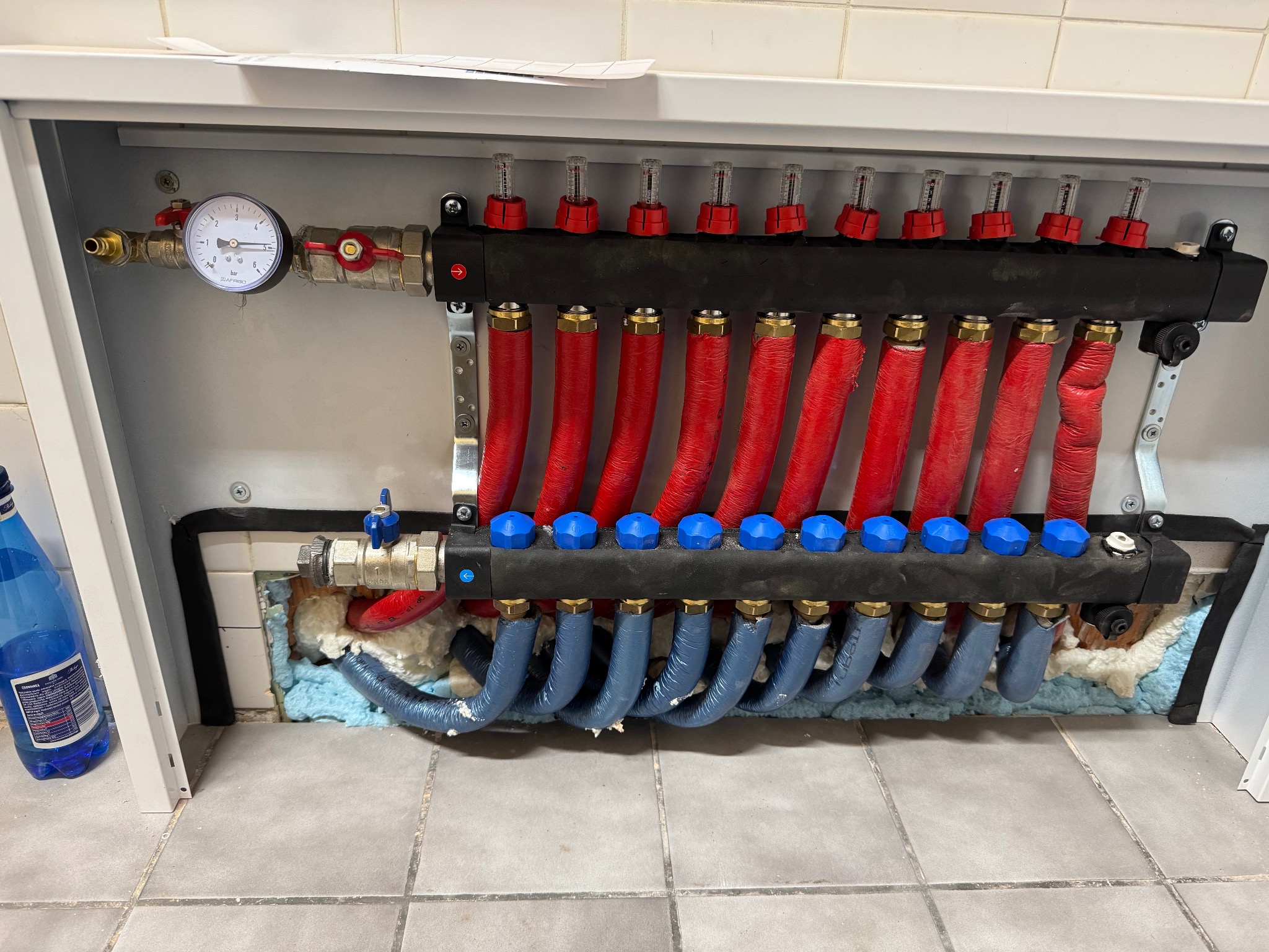 Rozdzielacz ogrzewania podłogowego w szafce, widoczne czerwone i niebieskie rury, manometr i zawory, montaż w trakcie uszczelniania pianką montażową.