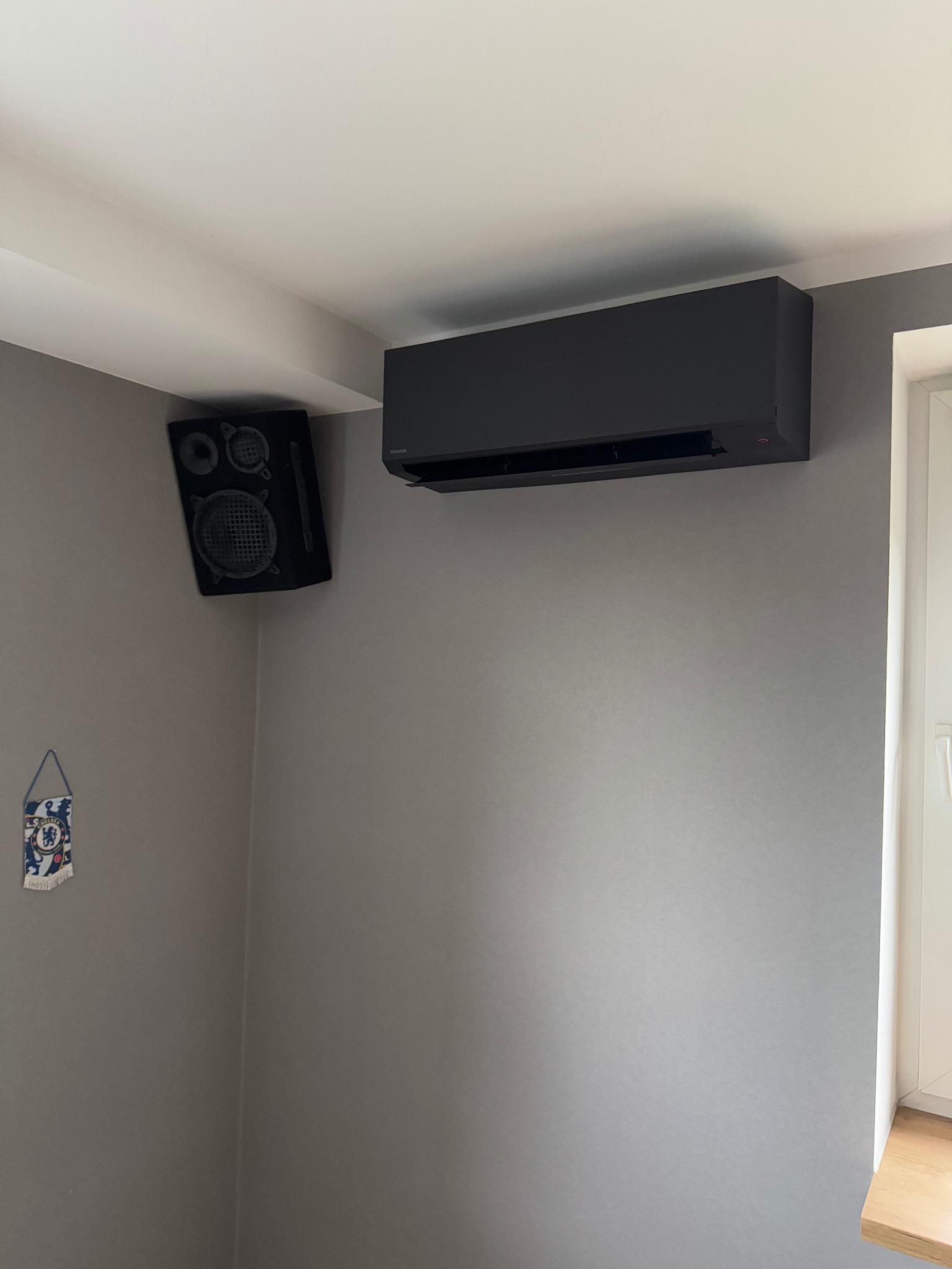 Czarny klimatyzator ścienny Toshiba, zamontowany w rogu pokoju z szarą ścianą, obok głośnika i małej flagi Chelsea.