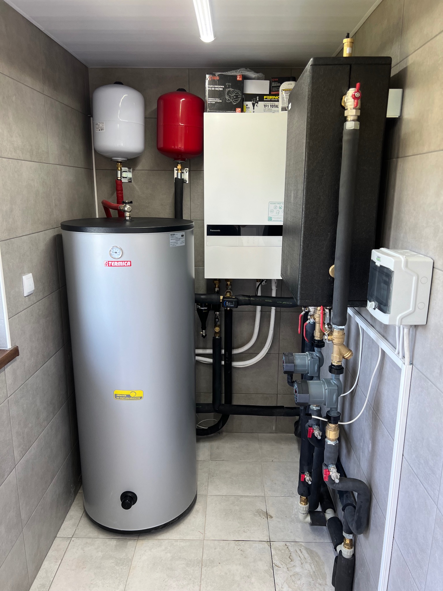 Kotłownia w szarych płytkach z białym kotłem, szarym zbiornikiem Termica, czerwonym i białym naczyniem przeponowym oraz czarnymi rurami z zaworami i pompami obiegowymi.