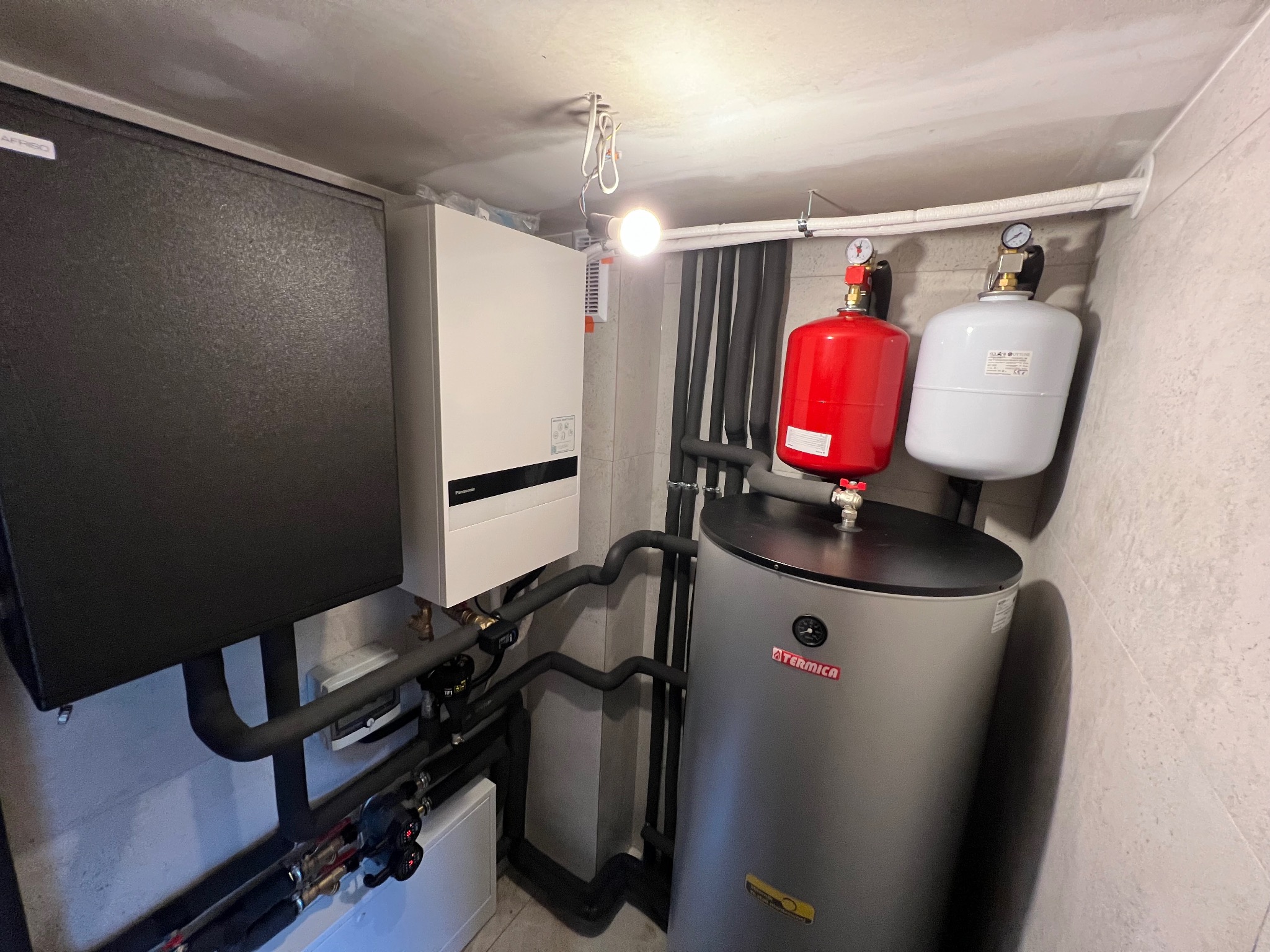 Kotłownia z zamontowaną pompą ciepła Panasonic Aquarea, zbiornikiem buforowym Termica i naczyniami przeponowymi w kolorach czerwonym i białym.