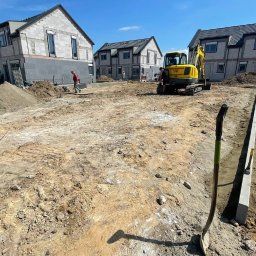 Marcin biuro - Żółta koparka na placu budowy z trzema surowymi domami z betonu komórkowego w tle, widoczna łopata wbita w ziemię i wytyczone krawężniki.