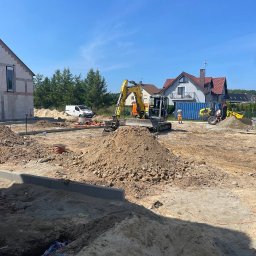 Marcin biuro - Przygotowanie terenu pod budowę domu: wykopane fundamenty, hałdy ziemi, mini koparki na placu budowy, widoczne rury kanalizacyjne i częściowo wzniesiony budynek w tle.