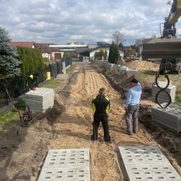 Marcin biuro - Przygotowanie podłoża i układanie betonowych płyt ażurowych na posesji, widoczny koparka, ubijarka i pracownicy w odblaskowych kamizelkach.