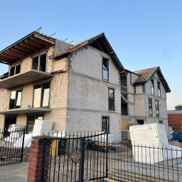 Marcin biuro - Niedokończony, dwupiętrowy dom z białego bloczka, widoczny zza czarnego, metalowego ogrodzenia. Widoczne materiały budowlane i elementy konstrukcyjne dachu.