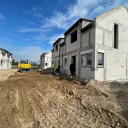 Budowa szeregowców z bloczków, widoczny mały żółty koparka i pracujący robotnicy. Częściowo zamontowane okna i orynnowanie. Niebo błękitne z chmurami.