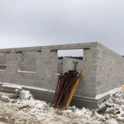 Ściany parteru wzniesione z bloczków betonu komórkowego na fundamencie, widoczne stemple budowlane i ośnieżone otoczenie. Budowa w trakcie zimy.