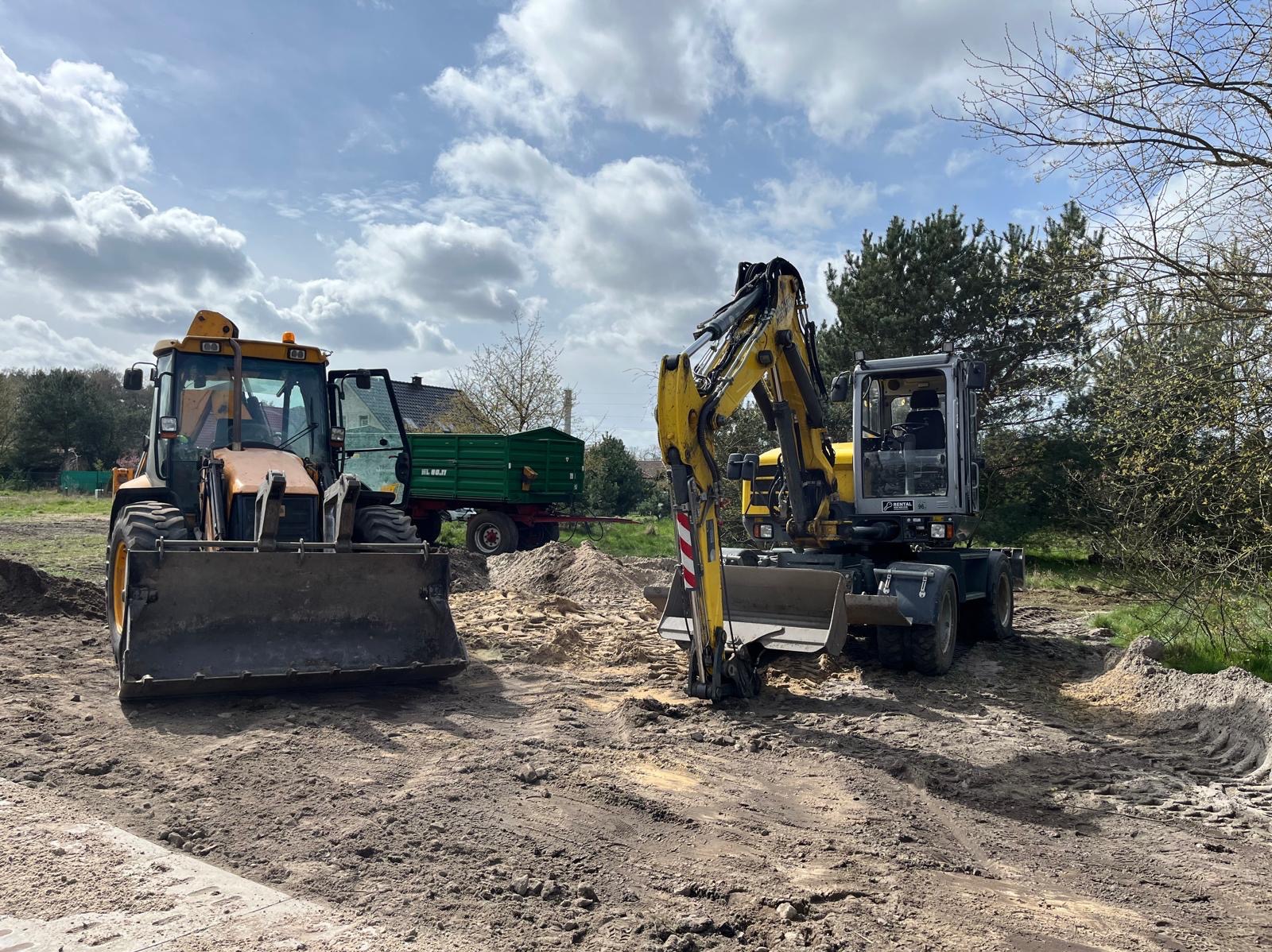 Żółto-czarny traktor z łyżką i koparka na placu budowy, w tle zielona przyczepa i budynek mieszkalny pod błękitnym niebem z chmurami.