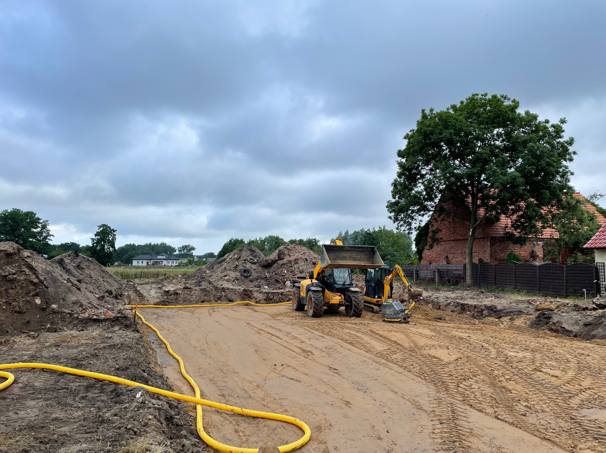 Żółty wąż na błotnistym terenie budowy z koparką i ładowarką na tle pochmurnego nieba i zabudowań.