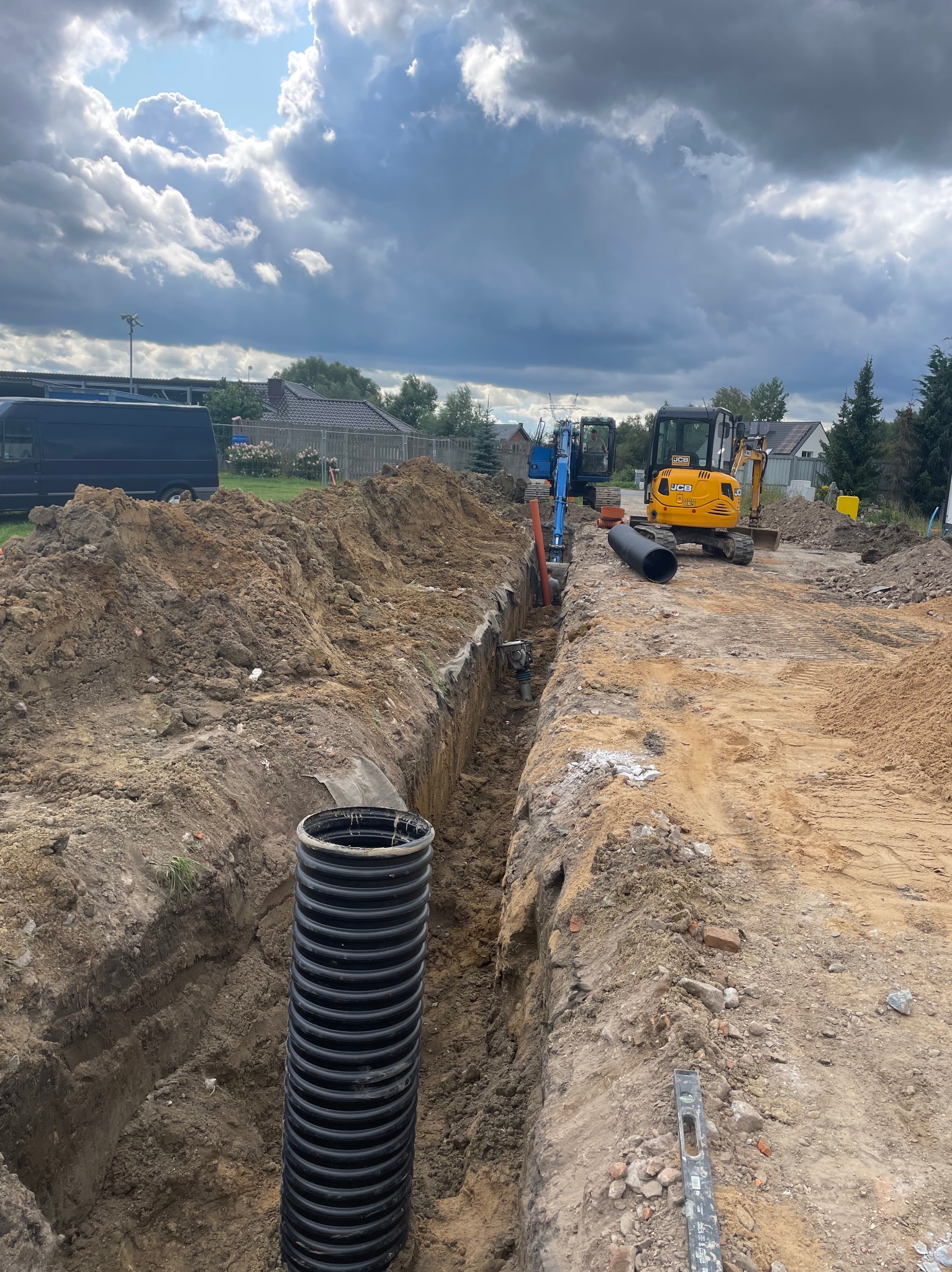Wykopany rów z widocznymi rurami kanalizacyjnymi i dwoma małymi koparkami JCB w tle, na placu budowy pod pochmurnym niebem.