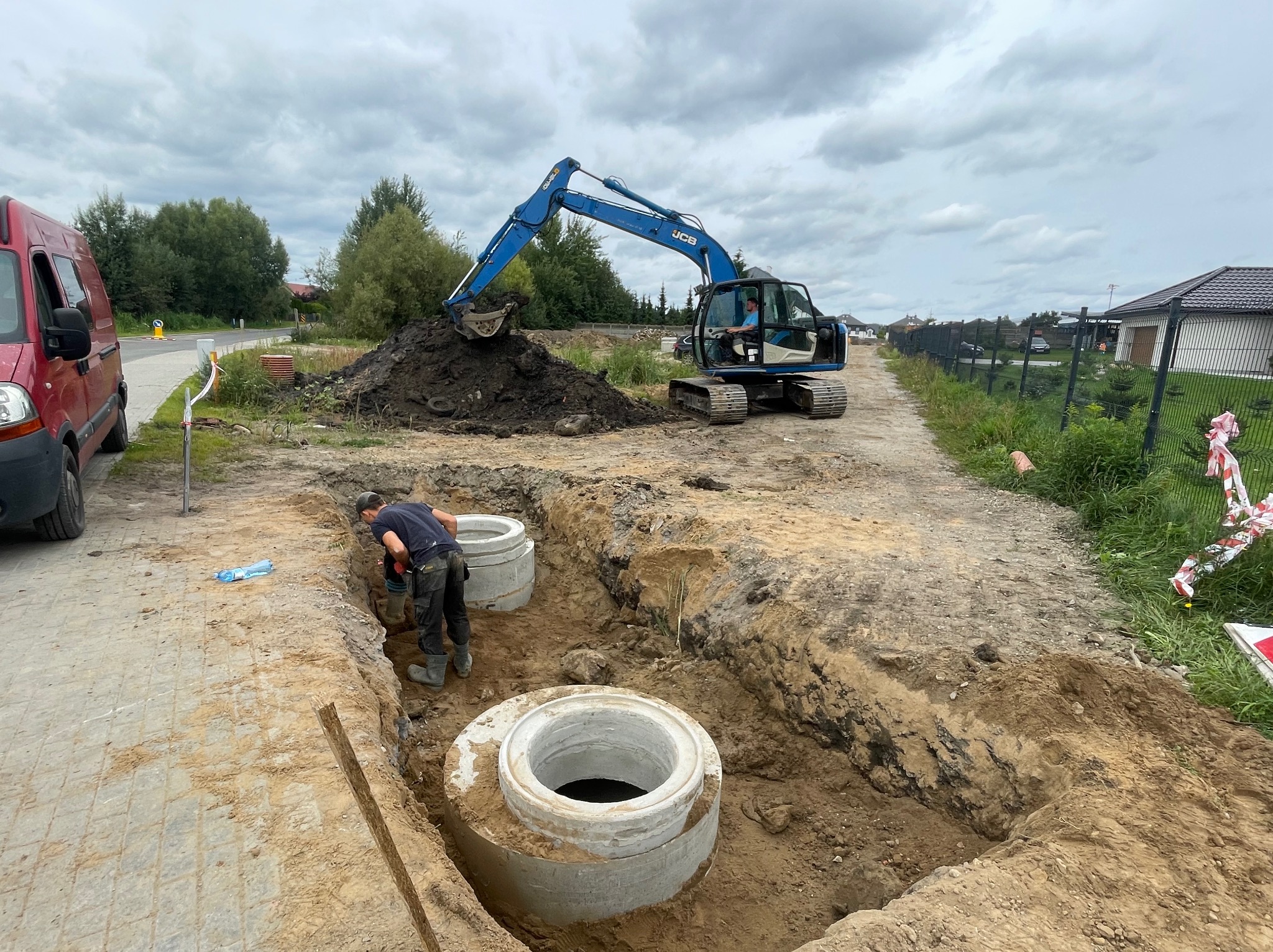 Pracownik w wykopie łączy betonowe kręgi szamba, w tle niebieska koparka JCB i sterta ziemi, obok stoi czerwony samochód dostawczy.