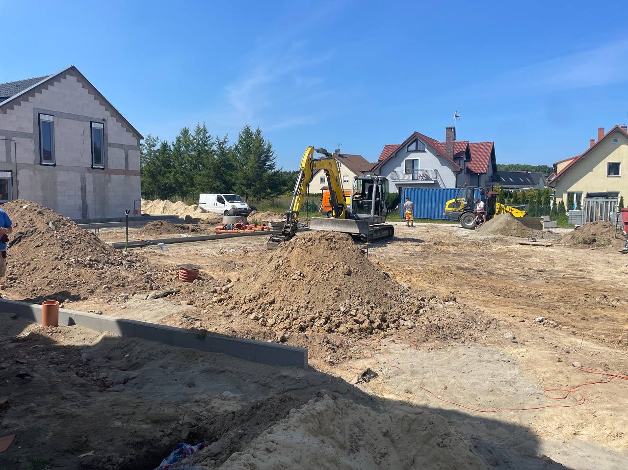 Przygotowanie terenu pod budowę domu: wykopane fundamenty, hałdy ziemi, mini koparki na placu budowy, widoczne rury kanalizacyjne i częściowo wzniesiony budynek w tle.