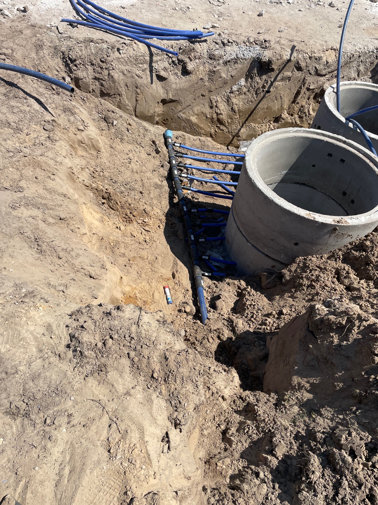 Wykop z ułożonymi rurami wodociągowymi i betonowymi kręgami kanalizacyjnymi, widoczny fragment gruntu i rur na powierzchni.