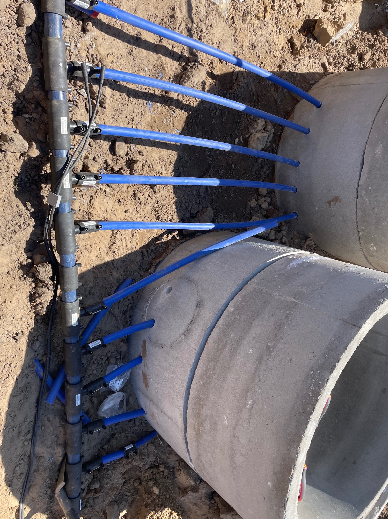 Instalacja niebieskich rur w betonowych kręgach w wykopie, widoczne elementy złączek i przewodów.