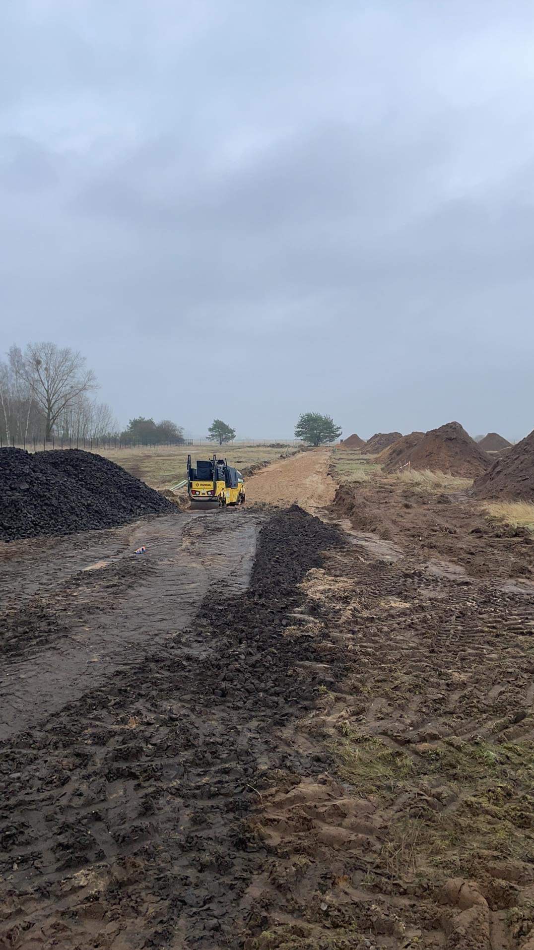 Żółty walec drogowy BOMAG ubija grunt pod budowę drogi, widoczne hałdy ziemi i kamieni, pochmurne niebo w tle.