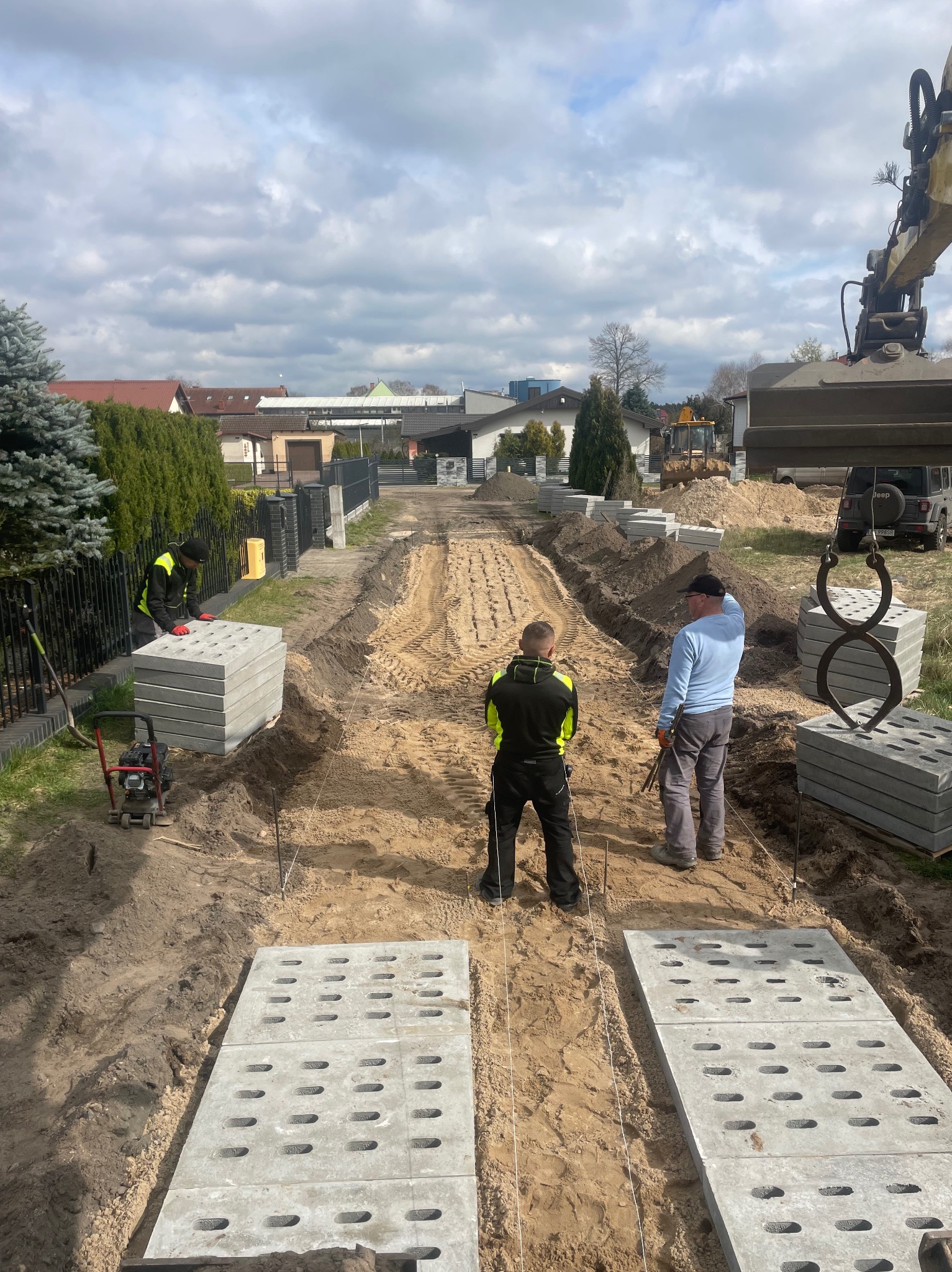 Przygotowanie podłoża i układanie betonowych płyt ażurowych na posesji, widoczny koparka, ubijarka i pracownicy w odblaskowych kamizelkach.