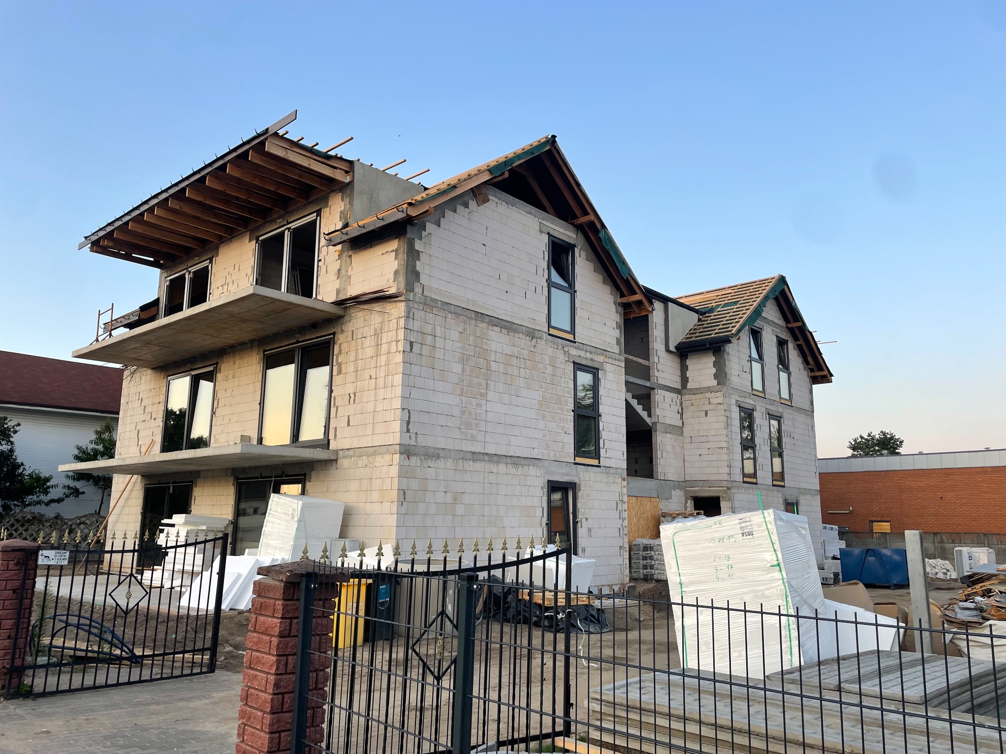 Niedokończony, dwupiętrowy dom z białego bloczka, widoczny zza czarnego, metalowego ogrodzenia. Widoczne materiały budowlane i elementy konstrukcyjne dachu.