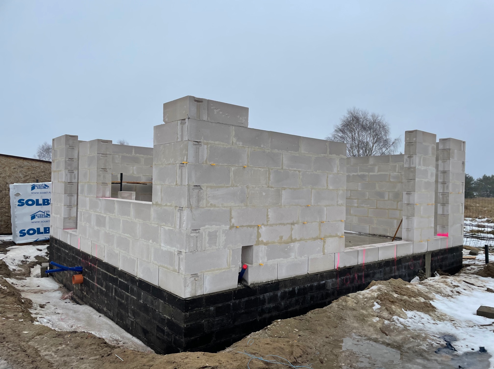 Wznoszenie ścian domu z bloczków SOLBET na fundamencie z izolacją przeciwwilgociową. Widoczne oznaczenia laserowe i fragmenty zbrojenia w tle. Zima.