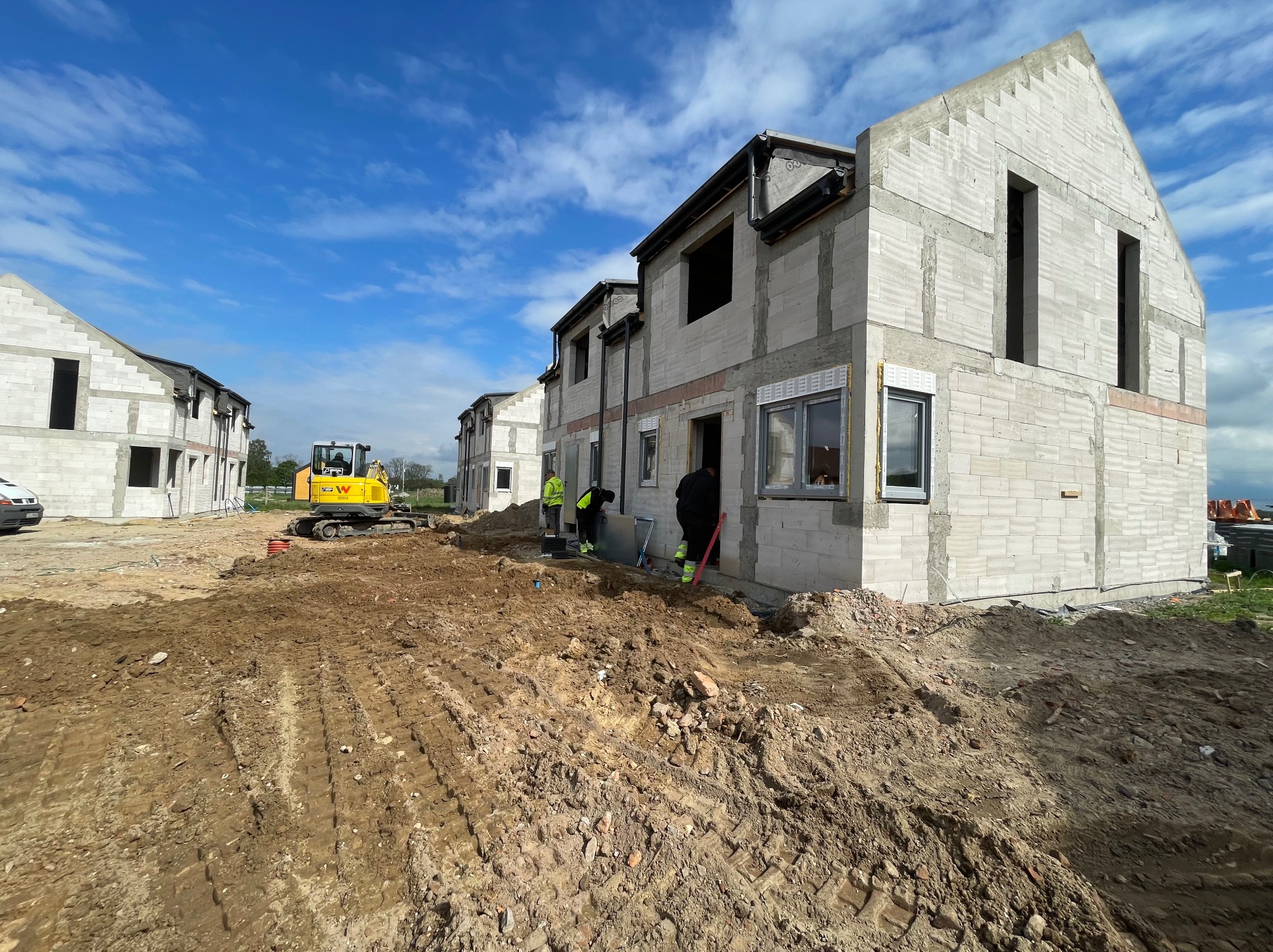 Budowa szeregowców z bloczków, widoczny mały żółty koparka i pracujący robotnicy. Częściowo zamontowane okna i orynnowanie. Niebo błękitne z chmurami.