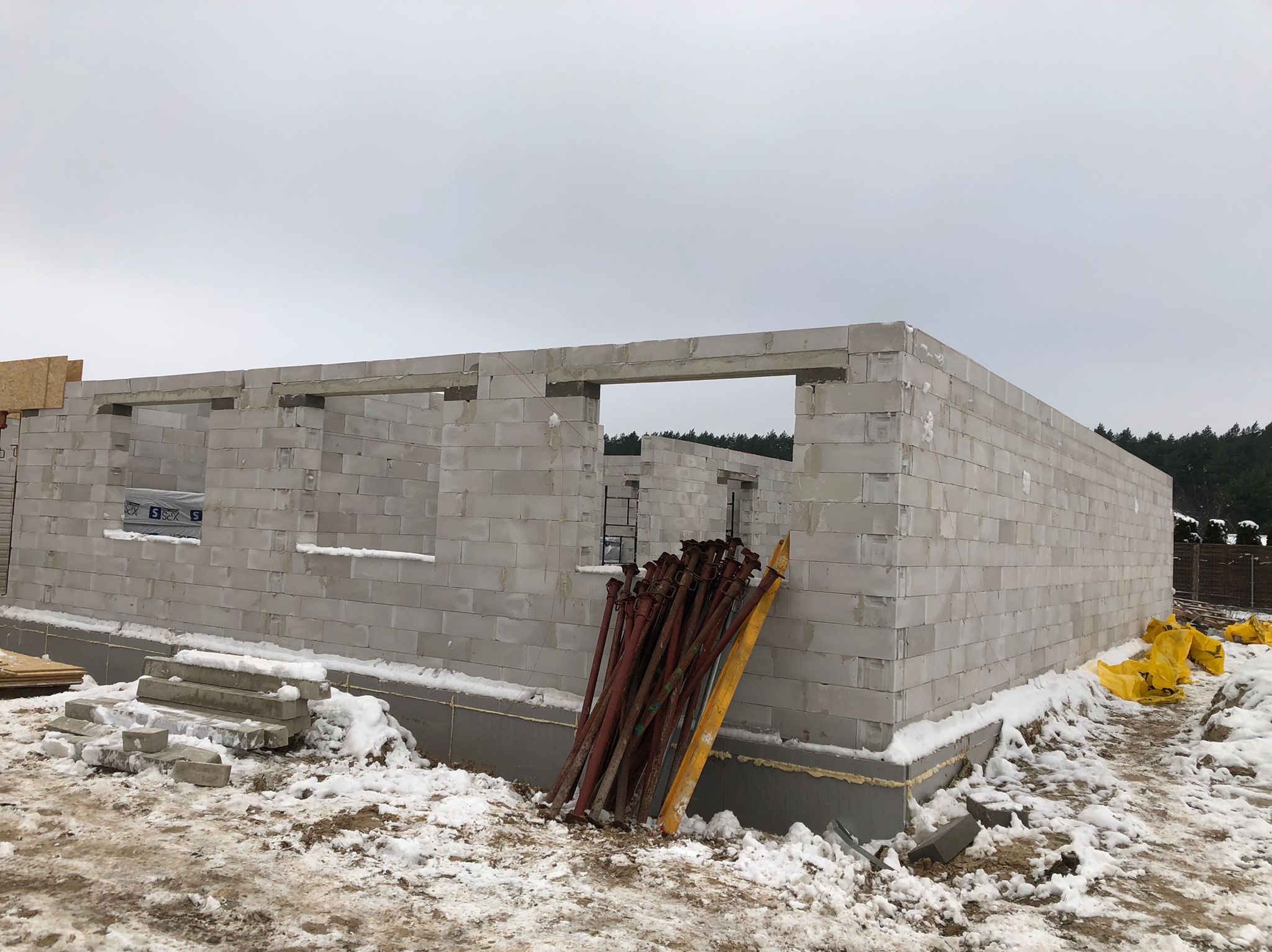 Ściany parteru wzniesione z bloczków betonu komórkowego na fundamencie, widoczne stemple budowlane i ośnieżone otoczenie. Budowa w trakcie zimy.