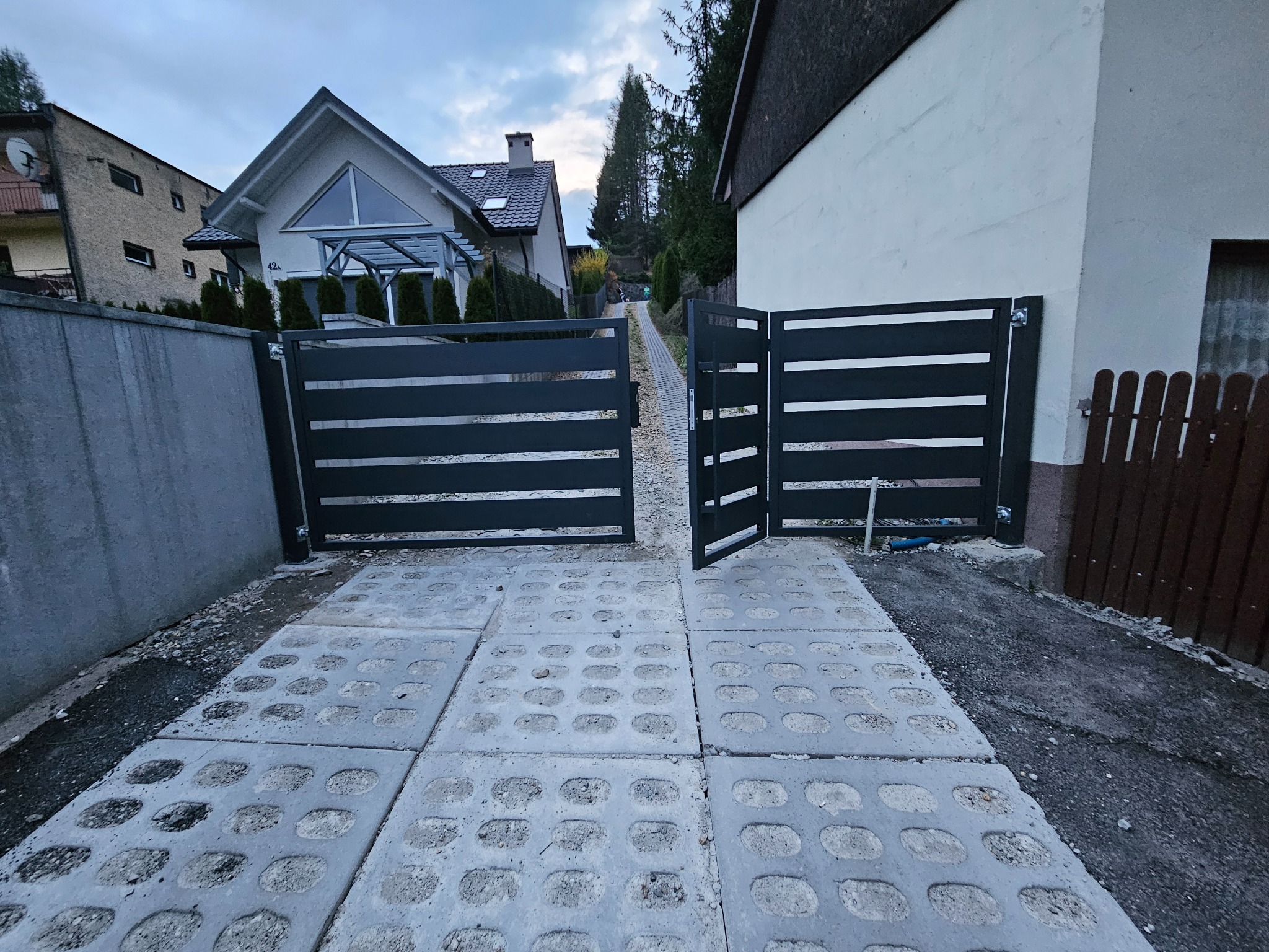 Nowoczesna, pozioma brama wjazdowa z metalowymi przęsłami na tle domu, widziana z perspektywy wjazdu wyłożonego ażurowymi płytami betonowymi.