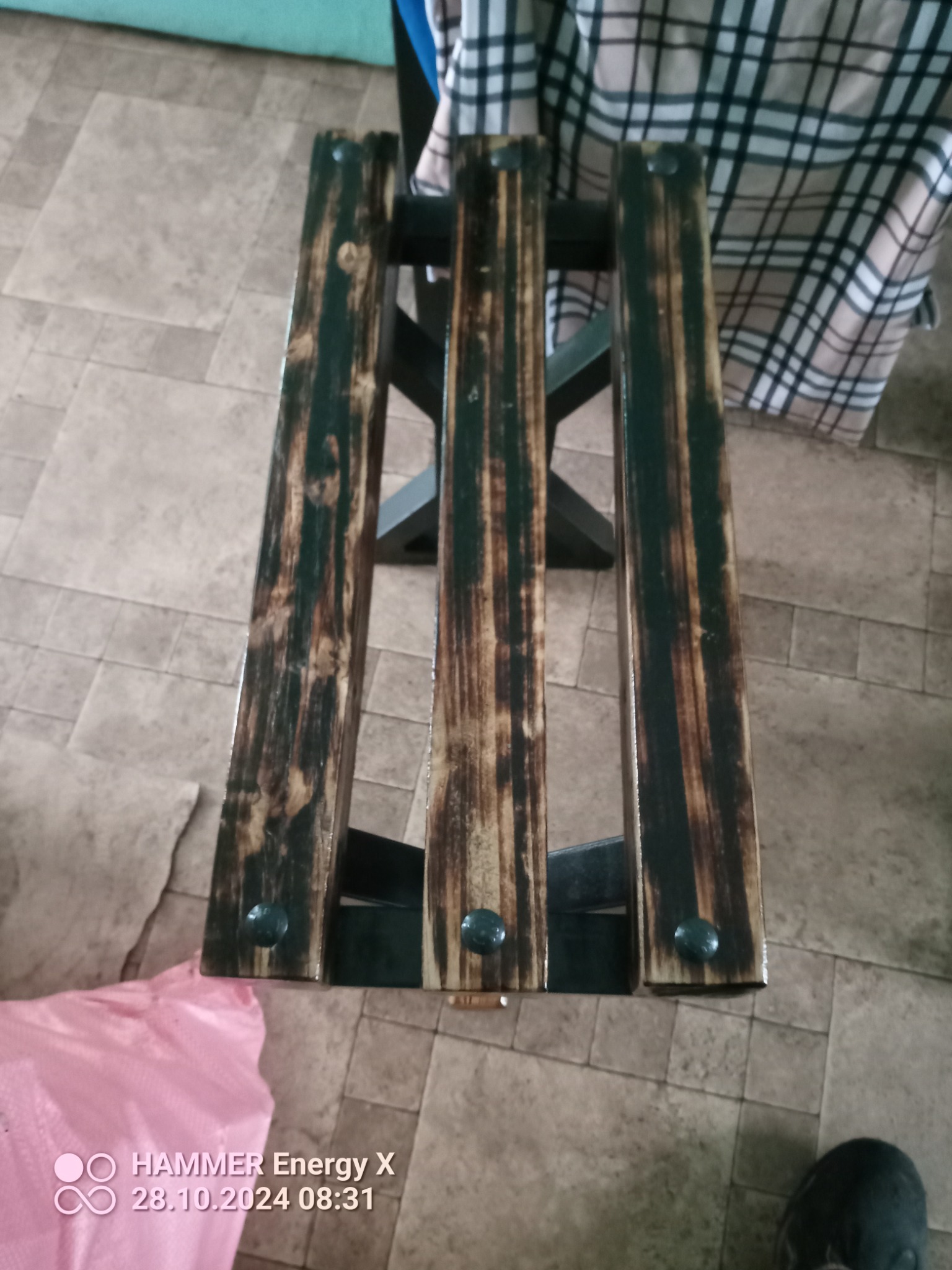 Drewniany taboret z trzema deskami i ciemnymi śrubami, z efektem opalania drewna, na tle podłogi z płytek i fragmentu ubrania w kratę.