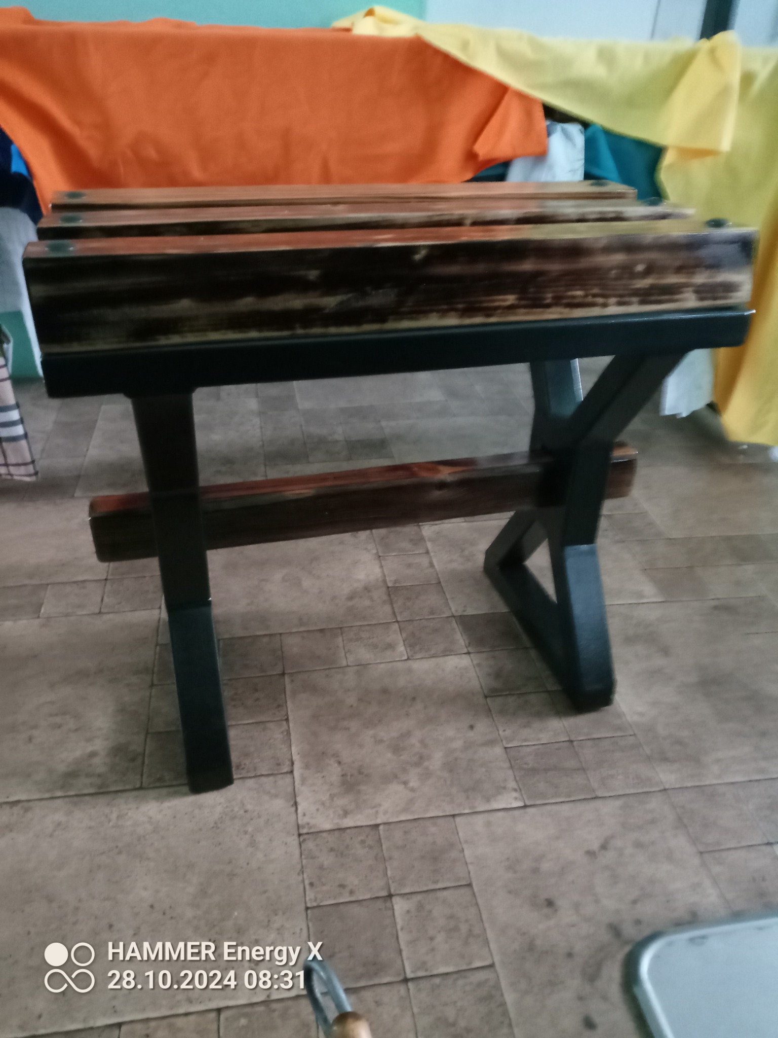 Stolik kawowy z drewnianym blatem i czarnymi, metalowymi nogami, wykonany ręcznie, na tle kolorowych tkanin.