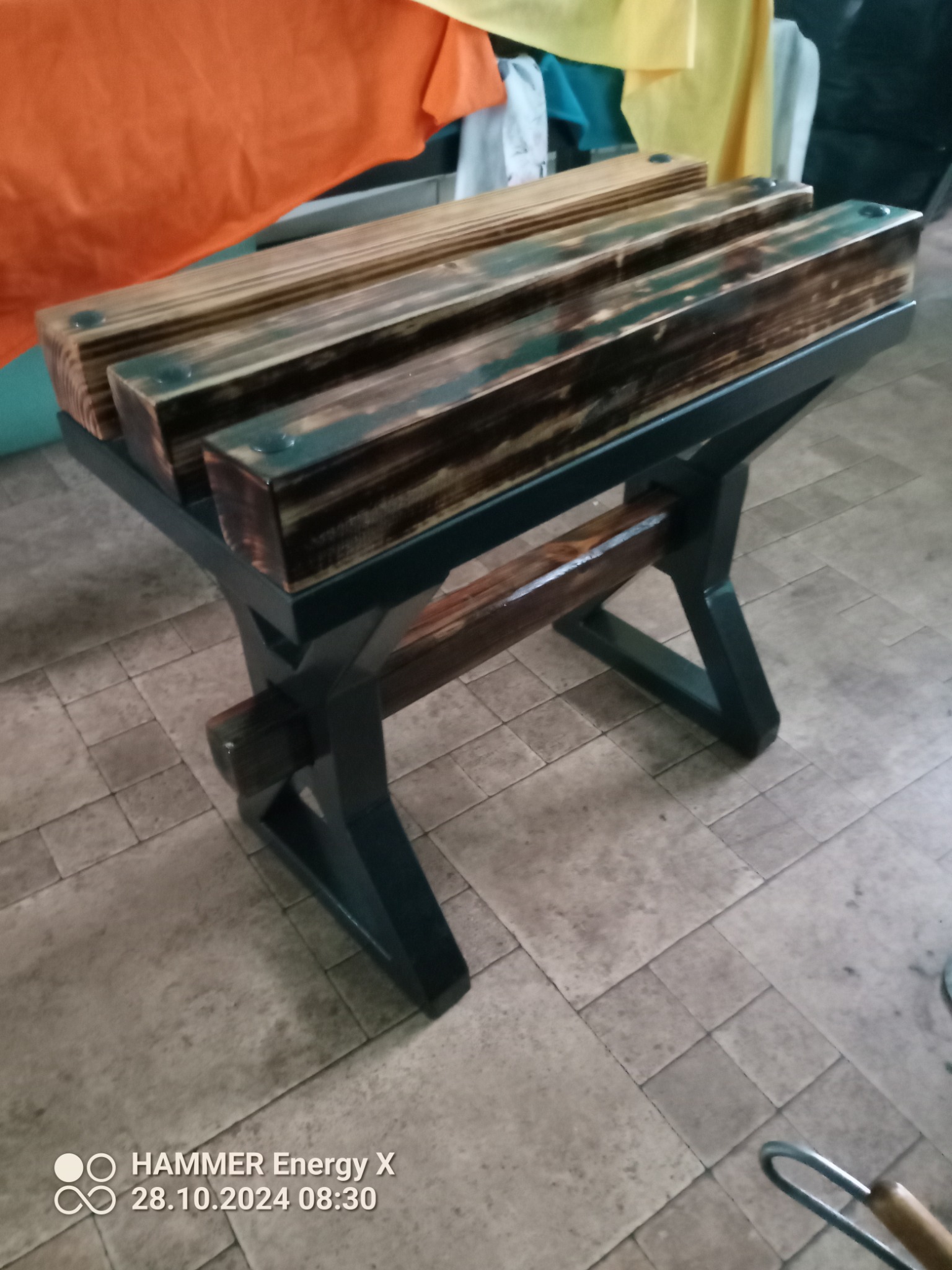 Stolik kawowy z metalową podstawą w kształcie litery X i drewnianym blatem z trzech belek, wykończony ciemną bejcą, widoczny na tle płytek podłogowych.