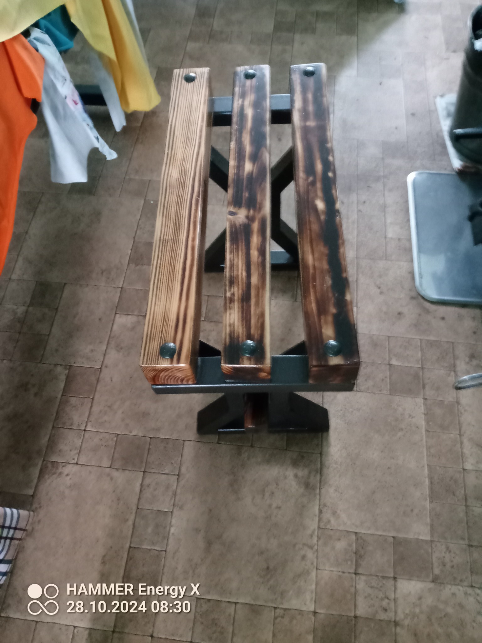 Metalowa konstrukcja spawana z trzema drewnianymi siedziskami, częściowo opalonymi, połączonymi nitami.