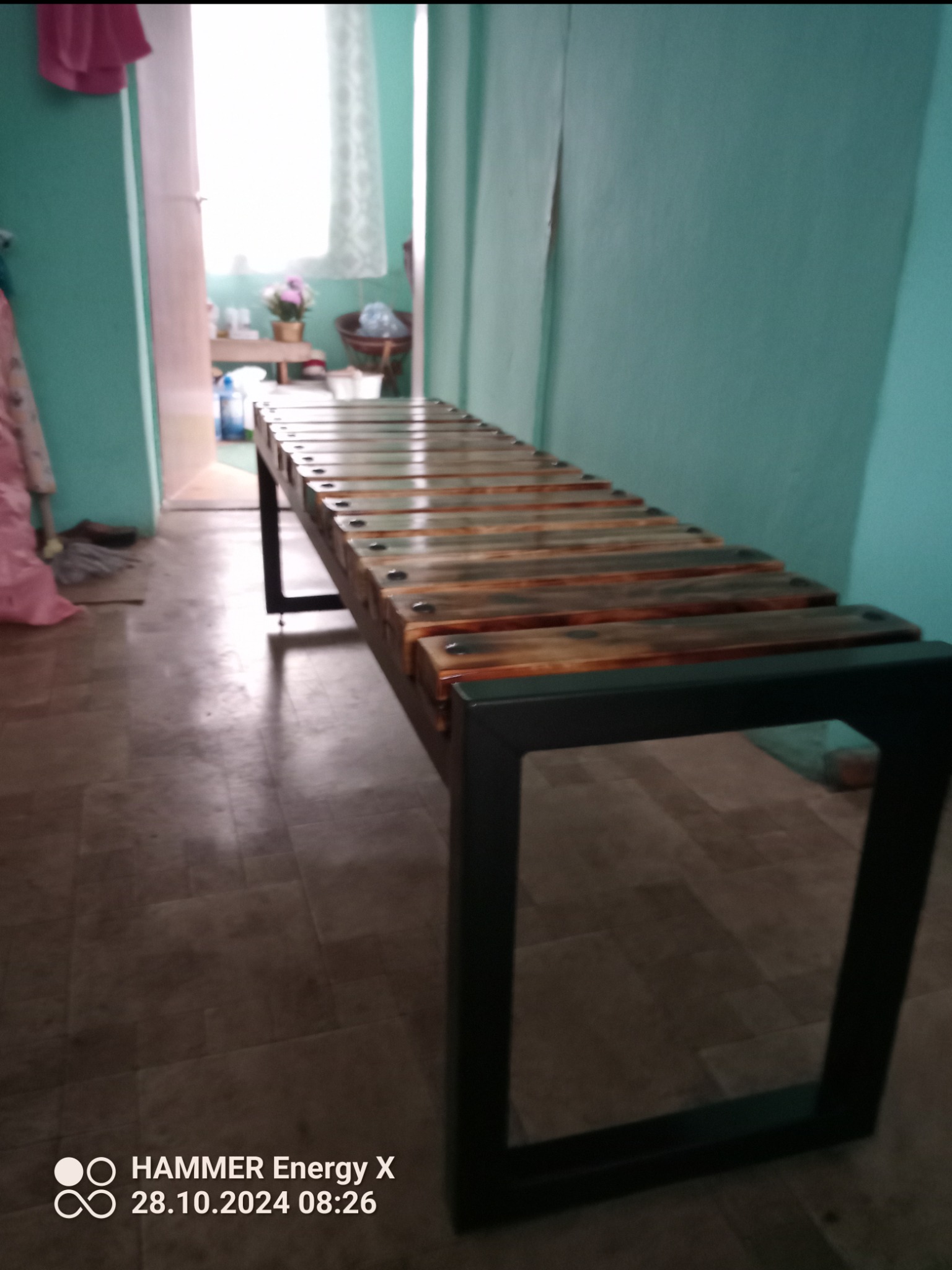 Metalowa konstrukcja ławki z drewnianymi szczebelkami, spawana z profili, widoczna w pomieszczeniu o turkusowych ścianach.