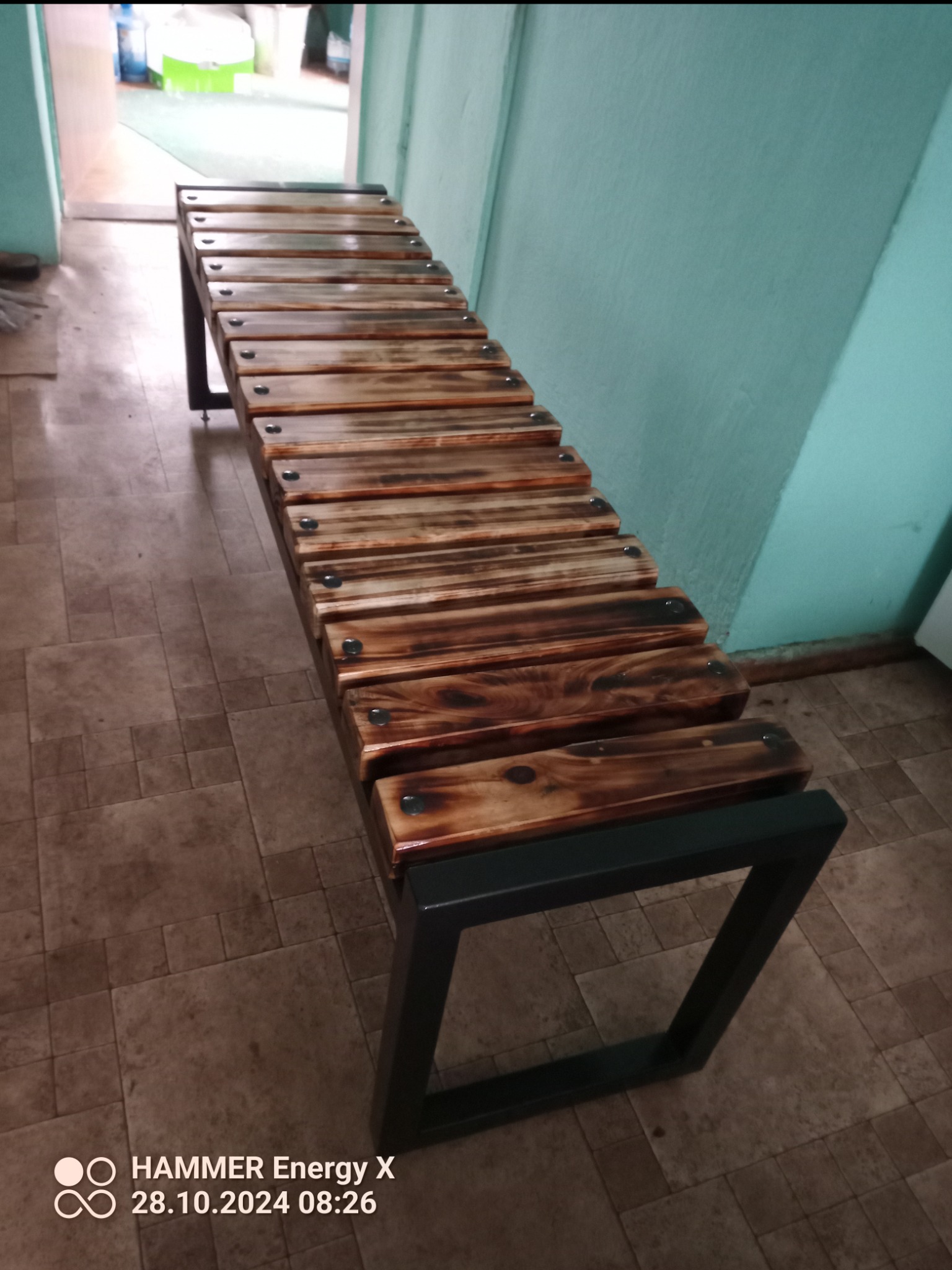 Metalowa ławka z drewnianymi szczeblami, opalana i lakierowana, na tle turkusowej ściany i korytarza.