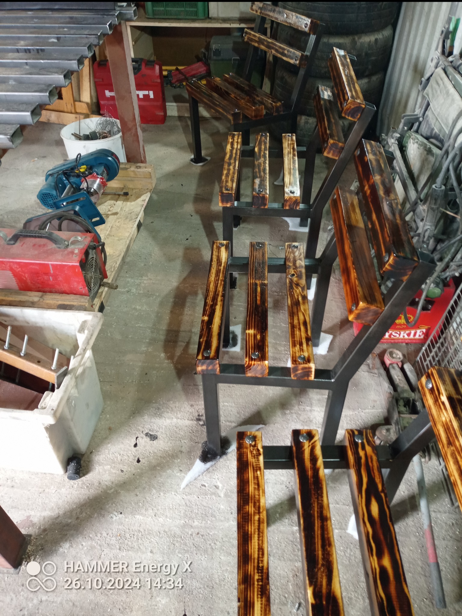 Metalowe stelaże z drewnianymi szczeblami, tworzące konstrukcję regału na tle warsztatu z narzędziami.