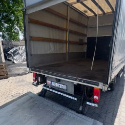 Transport- Przemysław Goździk - Otwarta paka busa z windą załadowczą, widok z tyłu. Na windzie czerwony znak z odciskiem stóp. Widoczna tablica rejestracyjna z Polski.