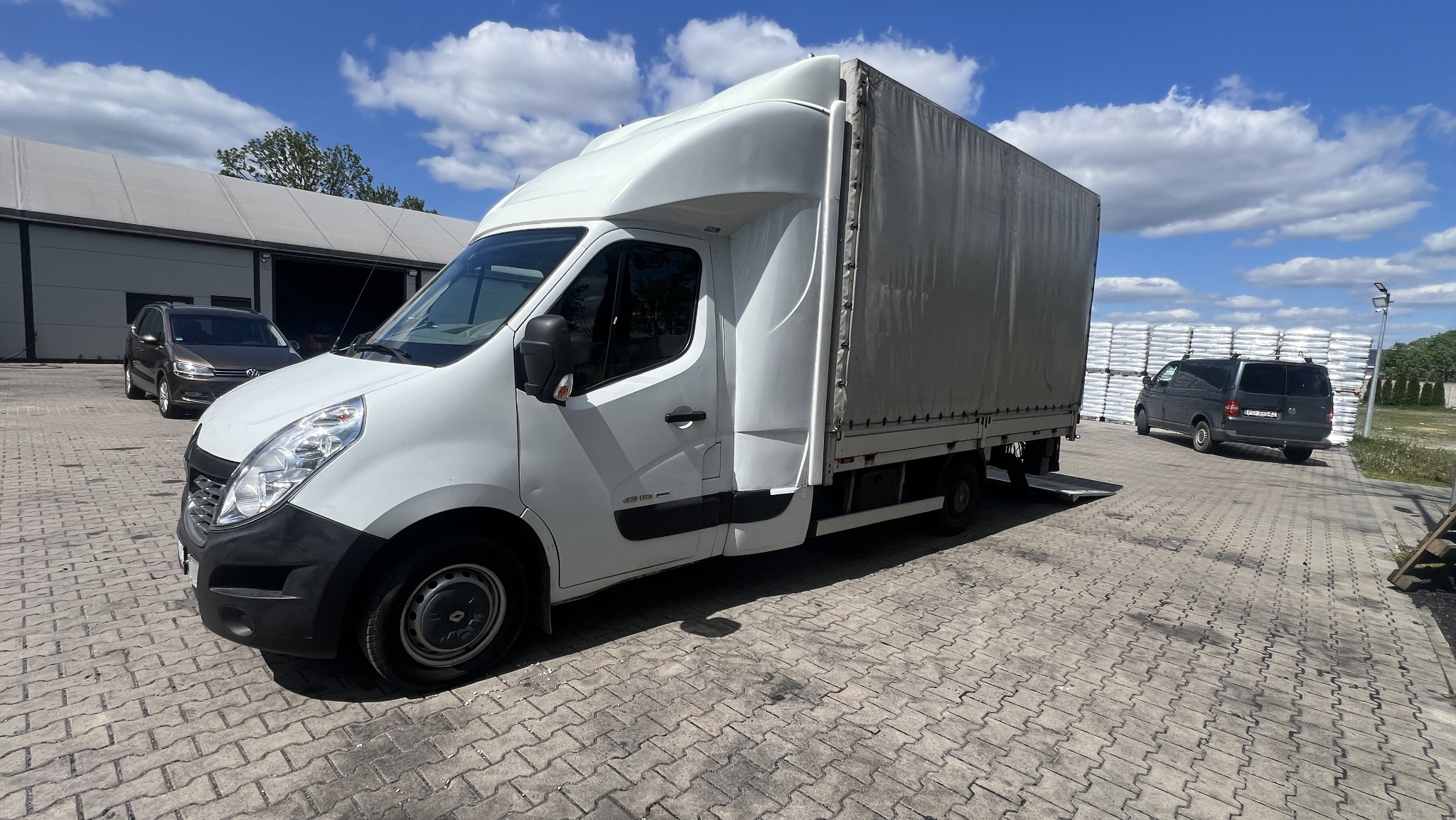 Biały bus z plandeką na brukowanym placu, w tle zaparkowany szary van i budynek magazynowy. Częściowo widoczne białe worki ułożone w stos.