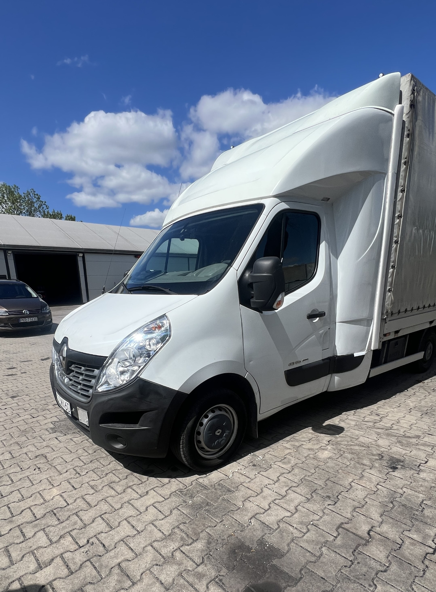 Biały Renault Master z plandeką na kostce brukowej, boczny widok. Na drugim planie budynek z bramami i zaparkowany samochód osobowy. Błękitne niebo z chmurami.