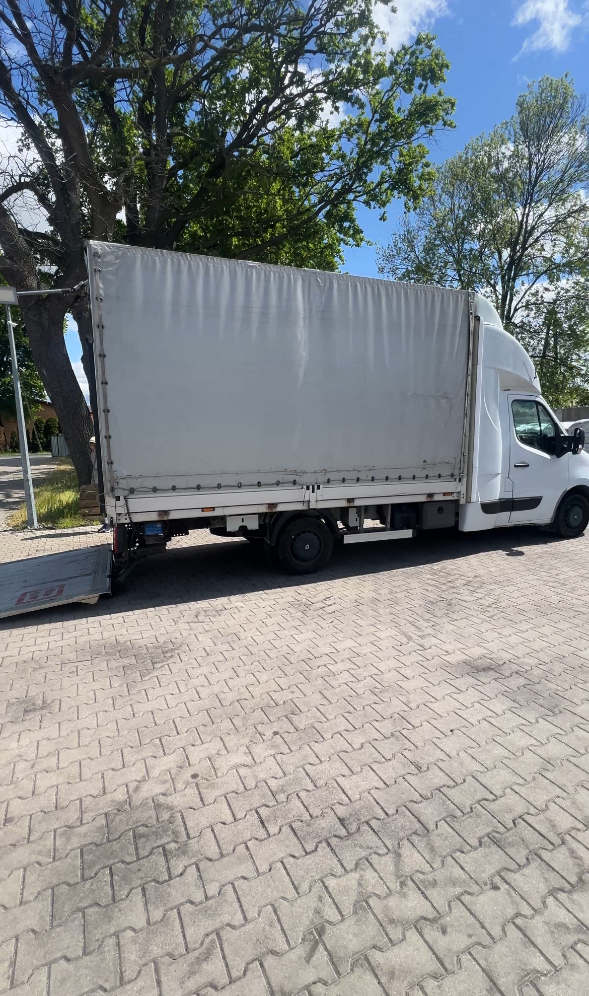 Biały bus z plandeką i opuszczoną rampą załadunkową, zaparkowany na brukowanym placu w słoneczny dzień. Widoczne drzewa w tle.