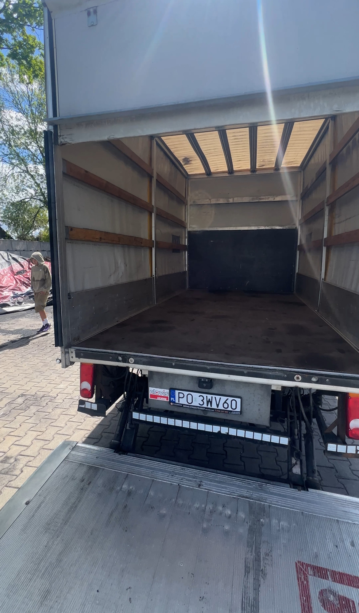 Pusta przestrzeń ładunkowa busa z otwartą klapą załadunkową. Widoczna tablica rejestracyjna i sylwetka osoby na zewnątrz. Dostępne światło dzienne.