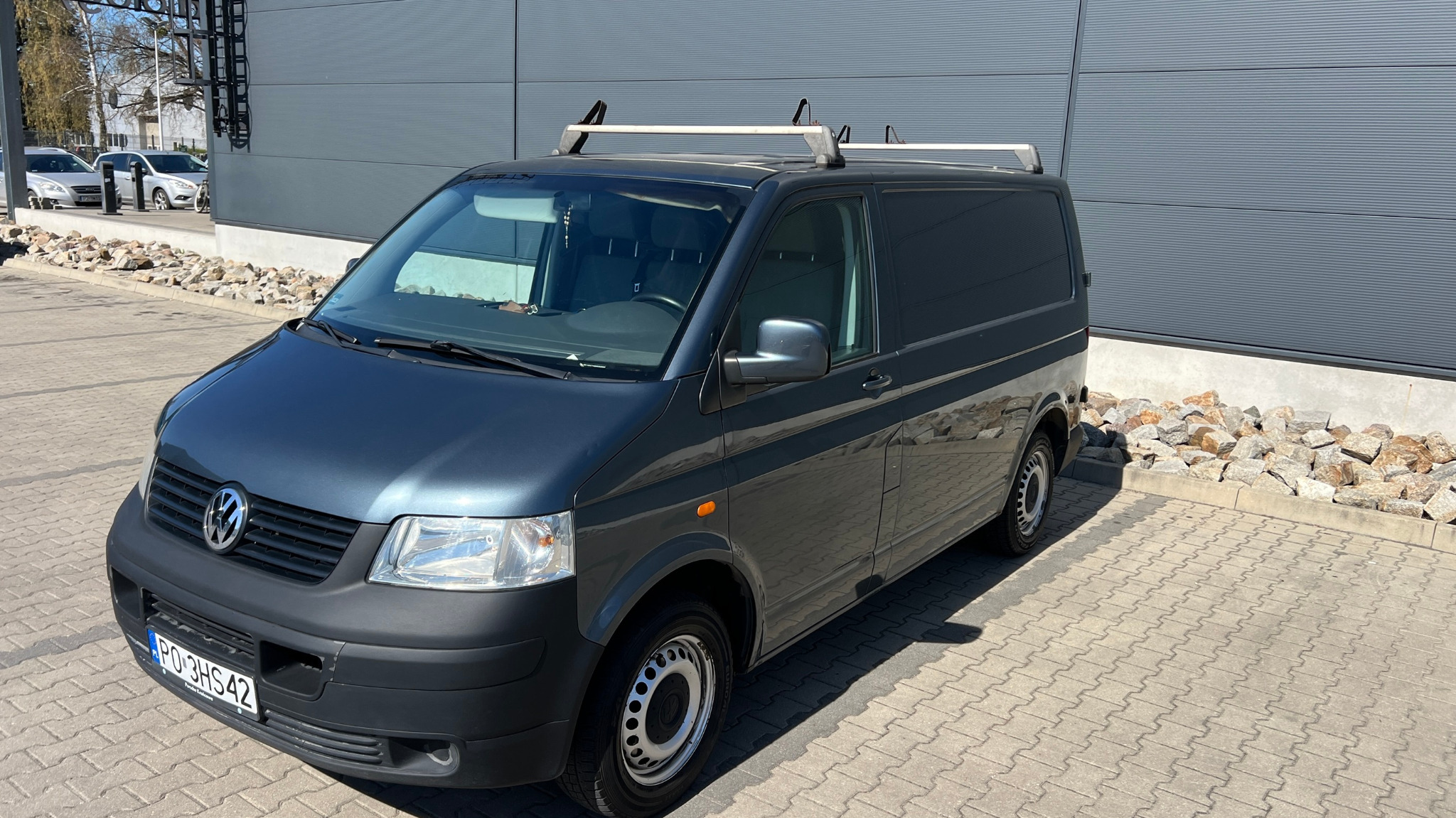 Szary Volkswagen Transporter T5 z relingami dachowymi, zaparkowany na kostce brukowej przed szarym budynkiem w słoneczny dzień. Widoczna tablica rejestracyjna i fragment otoczenia.