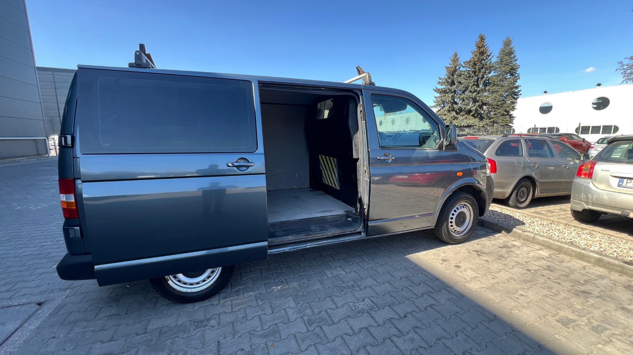 Szary bus z otwartymi drzwiami bocznymi na parkingu z kostki brukowej w słoneczny dzień. Widoczny bagażnik dachowy i inne zaparkowane pojazdy.