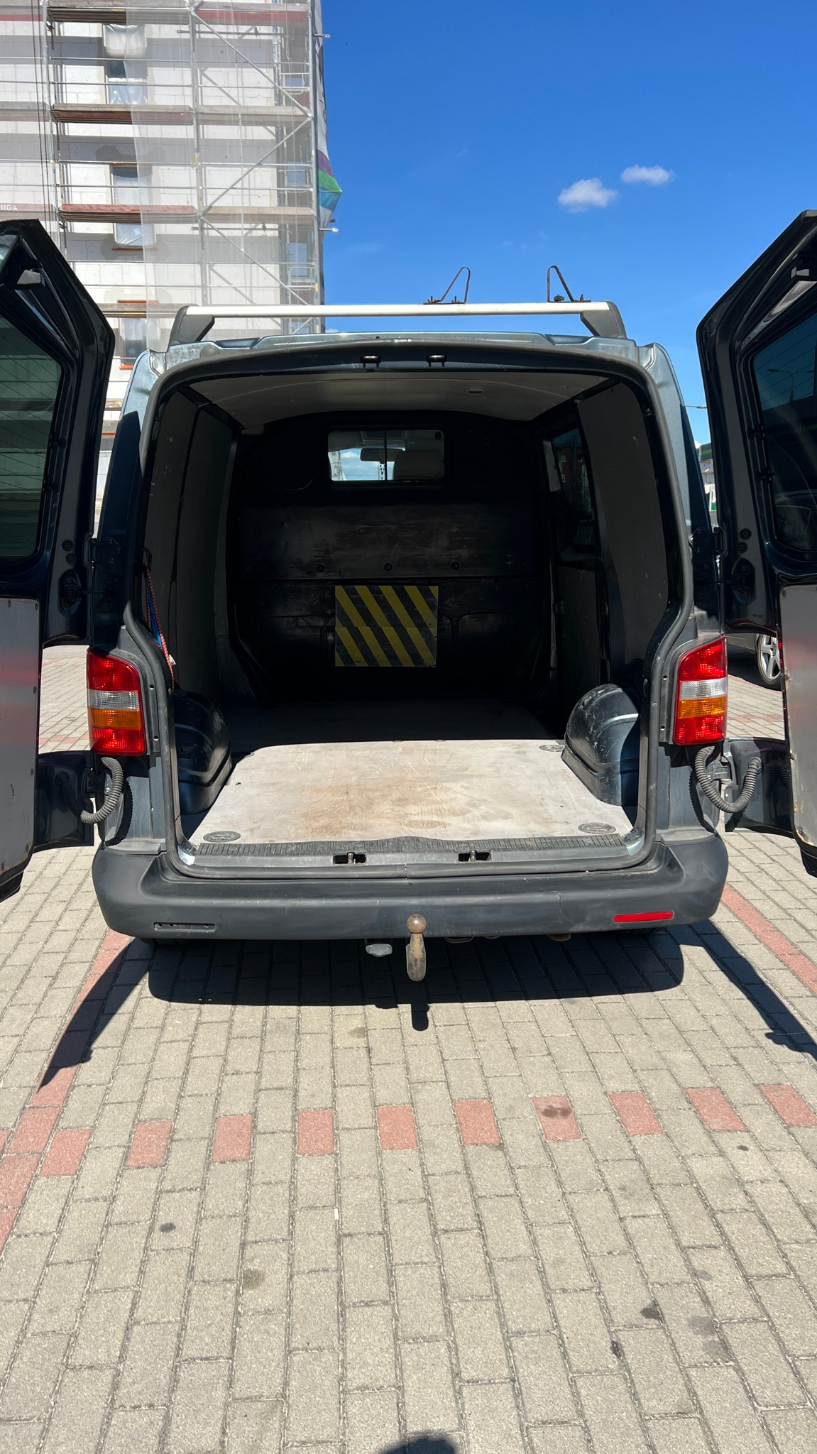 Otwarty, pusty bagażnik szarego busa z żółto-czarnym oznaczeniem na ścianie, na tle budynku w rusztowaniach i błękitnego nieba. Widok z zewnątrz na chodnik z kostki.