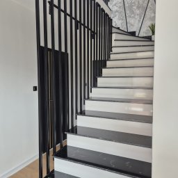 POZBRAM USŁUGI ŚLUSARSKO - SPAWALNICZE - PAWEŁ POZNAŃSKI - Nowoczesne schody z czarnymi, metalowymi poręczami i marmurowymi stopniami. Minimalistyczny design, jasne ściany i dekoracyjna faktura ściany w tle.