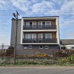 POZBRAM USŁUGI ŚLUSARSKO - SPAWALNICZE - PAWEŁ POZNAŃSKI - Dwukondygnacyjny dom z balkonami z poziomymi, szarymi barierkami. W tle błękitne niebo z chmurami. Na pierwszym planie metalowe ogrodzenie posesji.