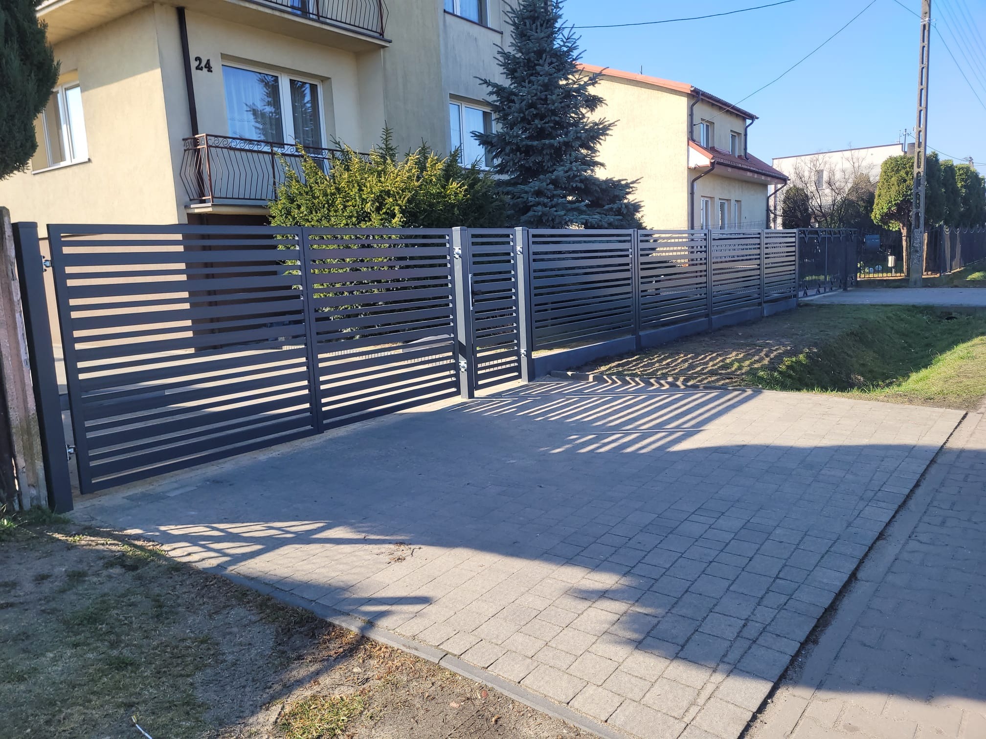 Nowoczesne, poziome ogrodzenie panelowe w kolorze grafitowym, z bramą wjazdową, na tle domu z numerem 24 i zielenią. Słoneczny dzień.
